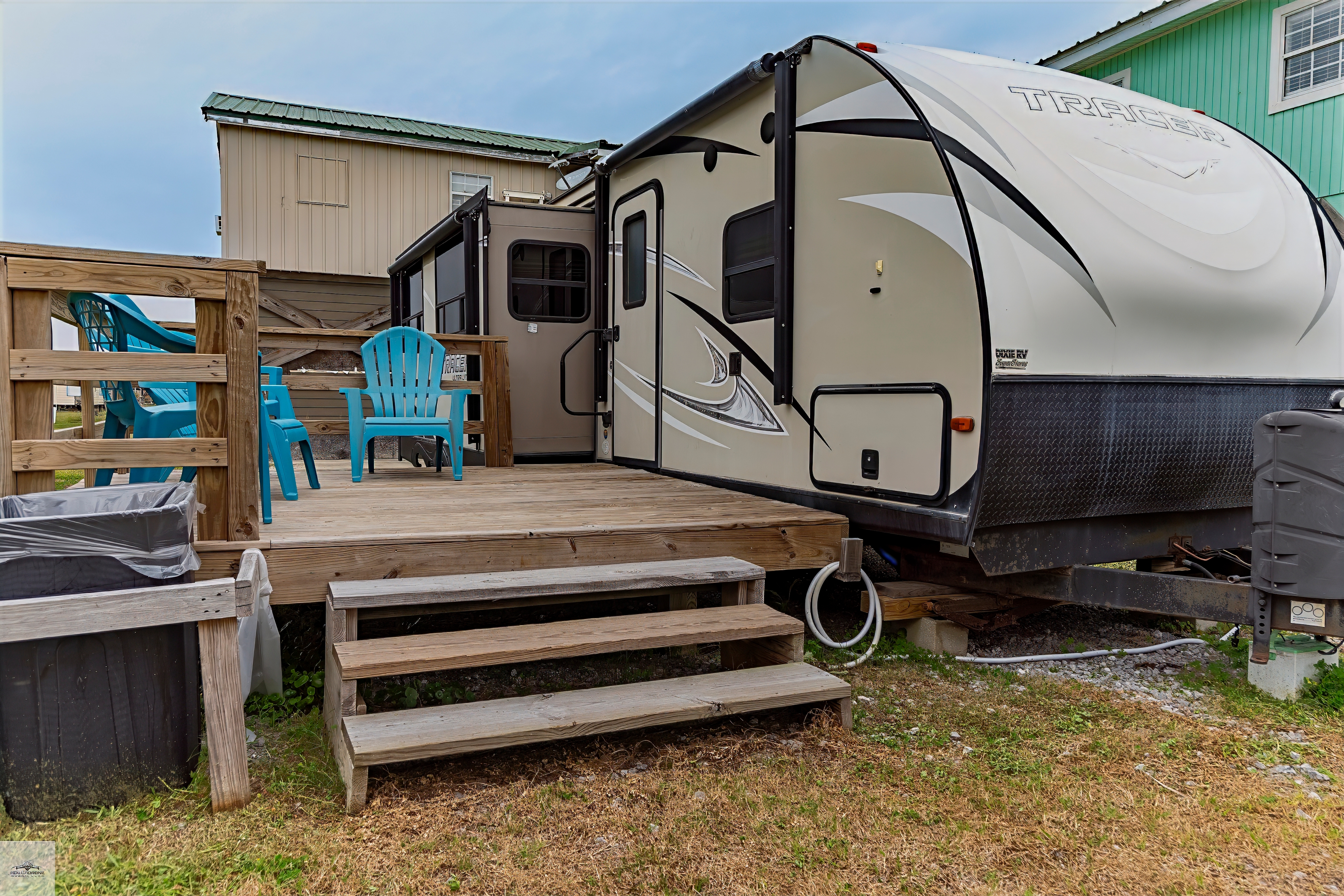 An RV/travel trailer parked on a grassy lot, featuring a small wooden deck with steps and a blue Adirondack chair outside the entrance.