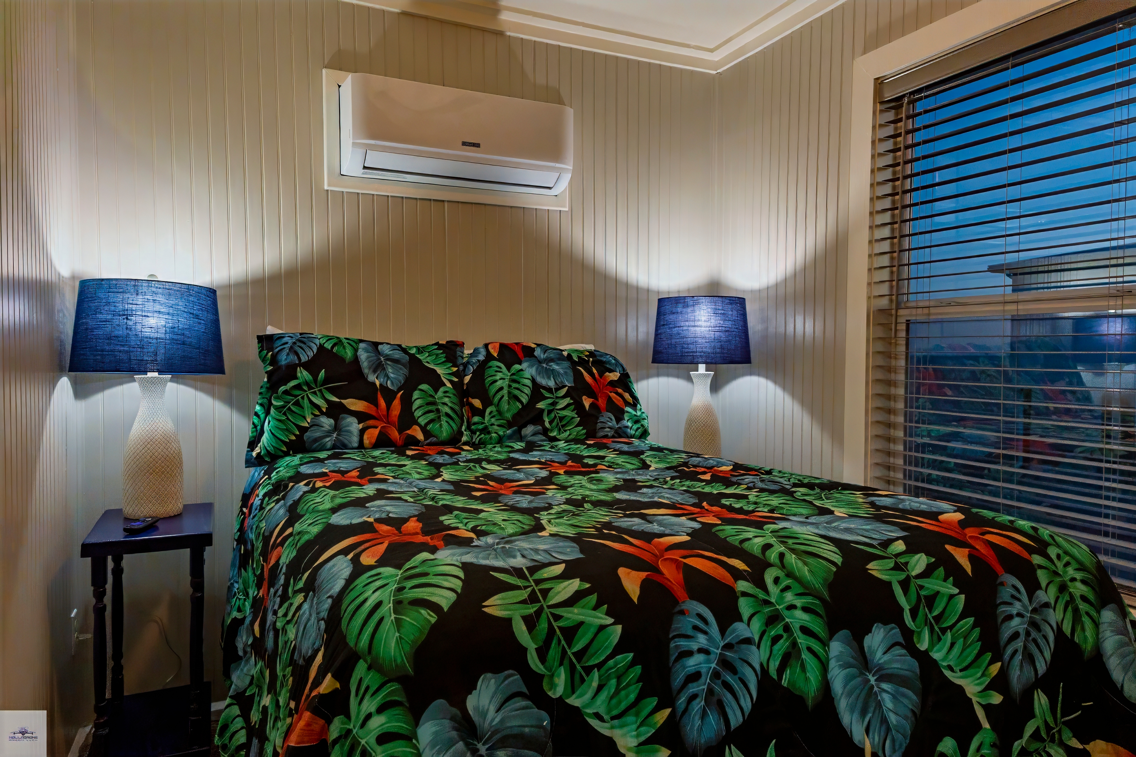 A close-up of a bedroom with a bed featuring a tropical floral bedspread, flanked by two matching lamps, and an AC unit above.