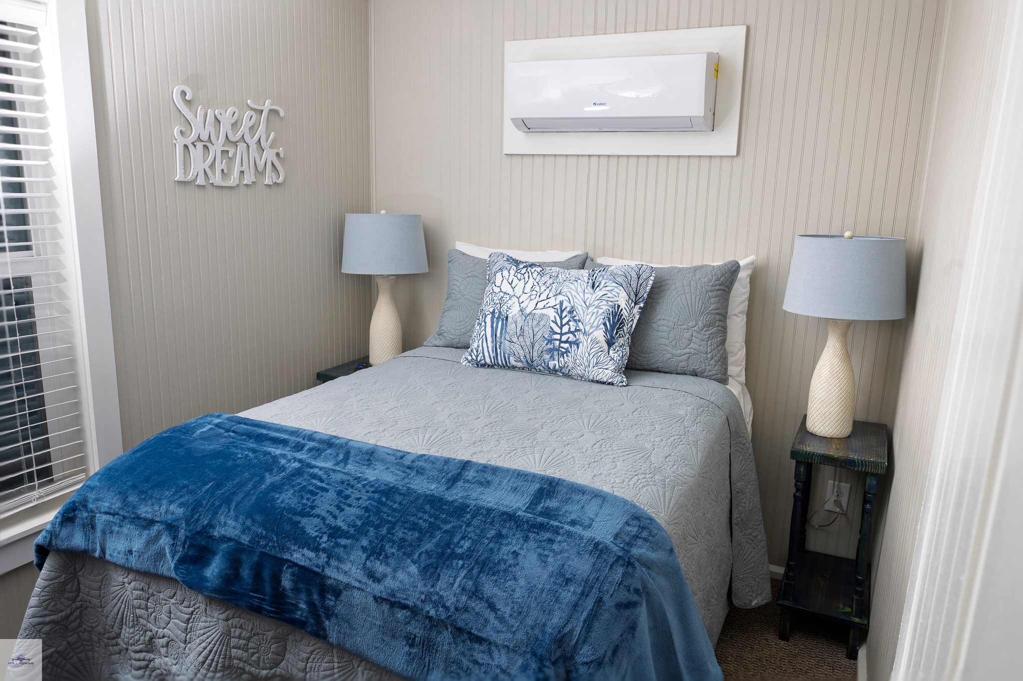 A close-up of a bedroom with a bed featuring a blue and grey bedspread, flanked by two matching lamps, and an AC unit above.