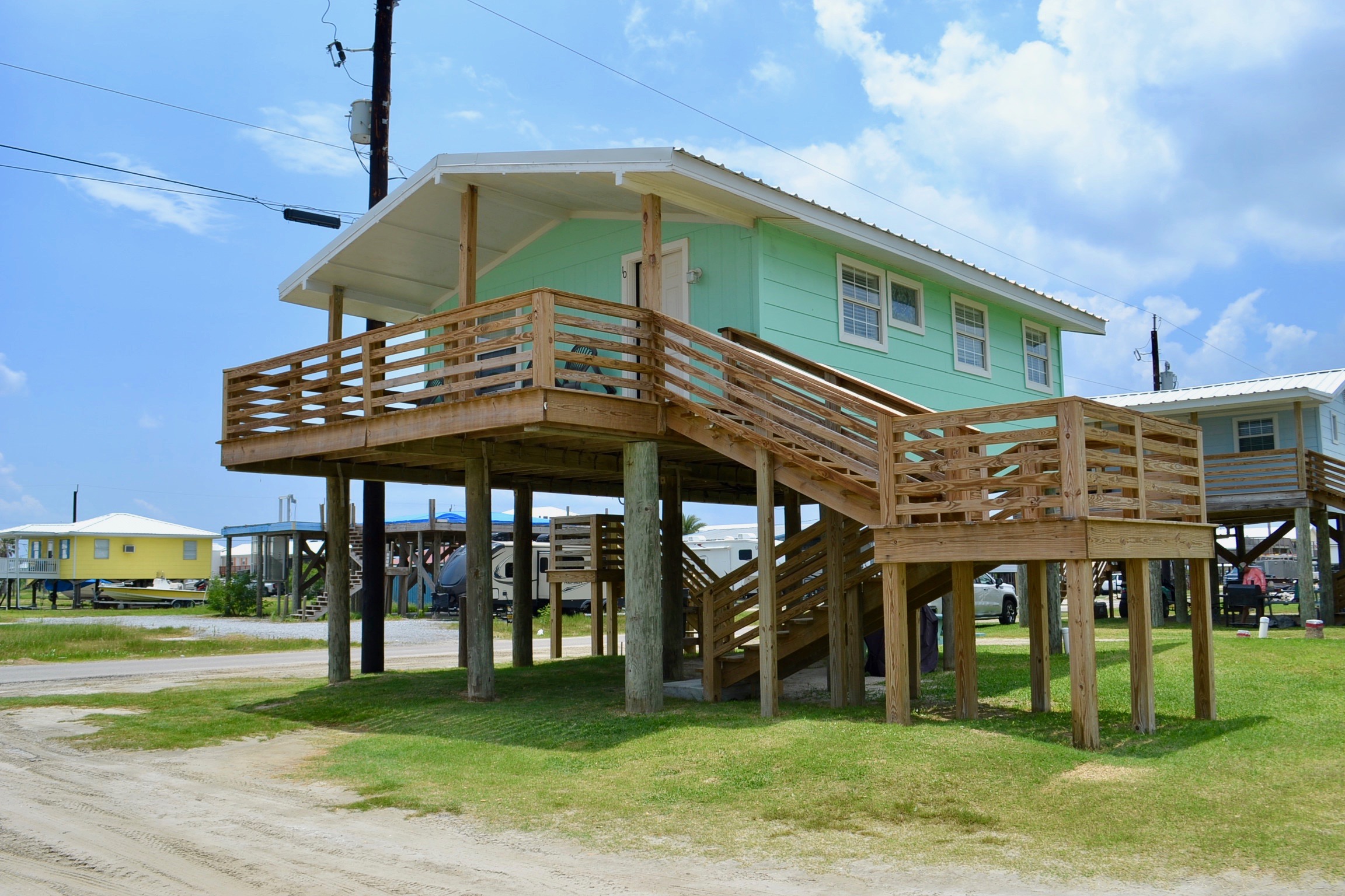 A charming, elevated stilt cottage painted mint green with a white trim, featuring a large wooden deck, stairs, and a covered porch.