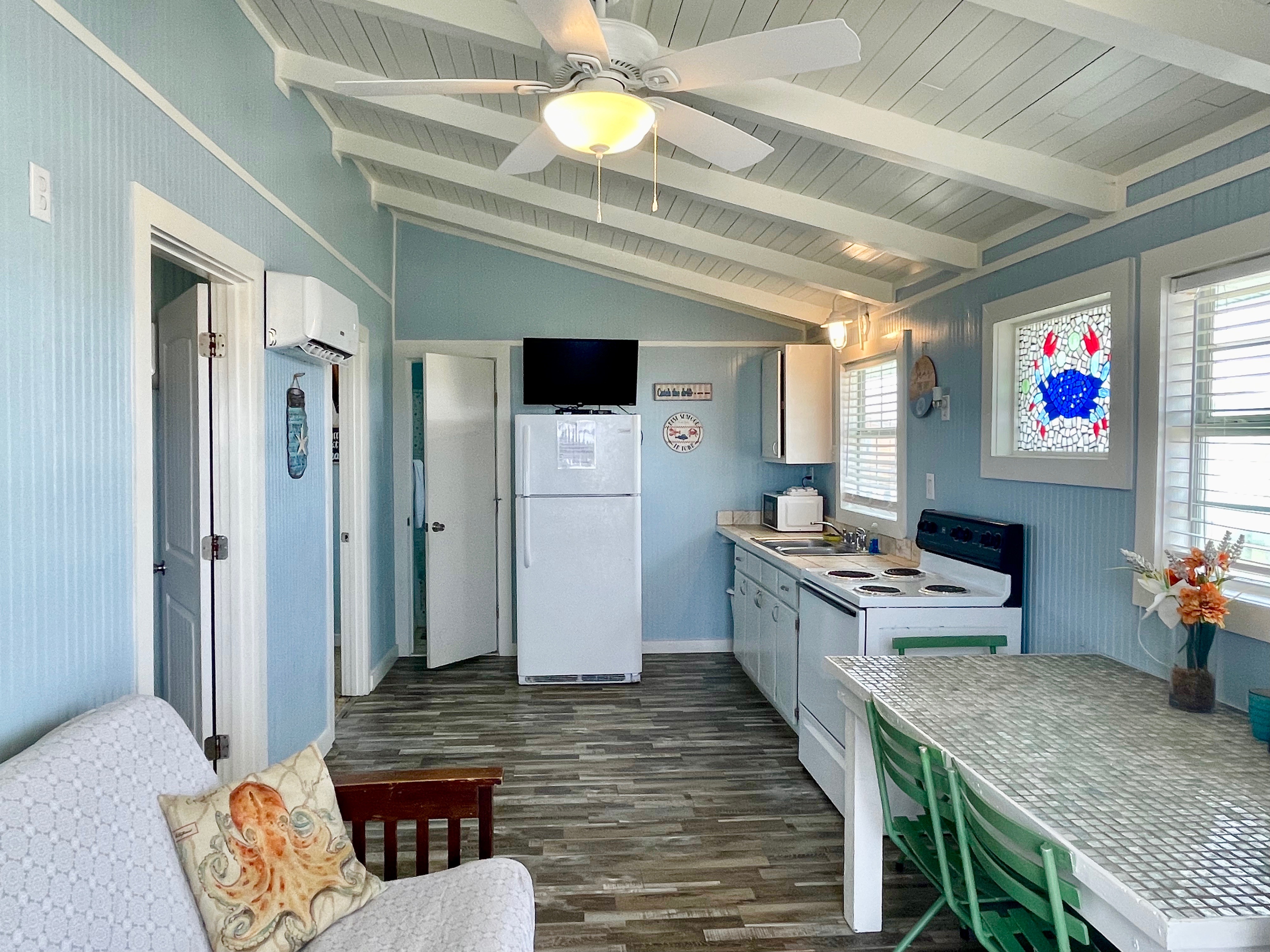 A brightly lit, coastal-themed cottage interior with light blue walls, a sloped white ceiling, a compact kitchen, and a white fridge.