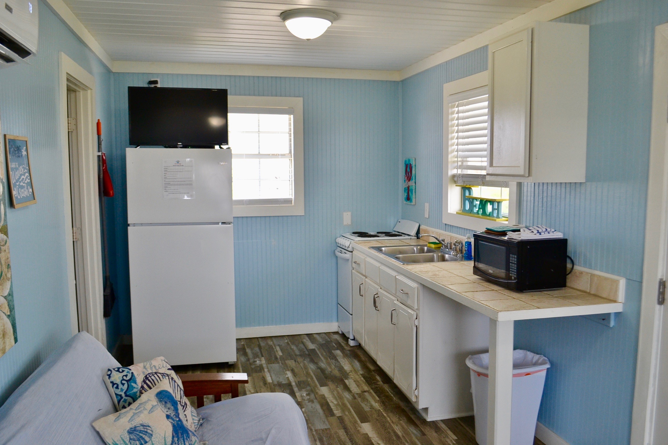 A bright kitchenette with light blue walls, featuring a white fridge with a TV mounted above it, white cabinets, and a futon slightly visible in foreground..