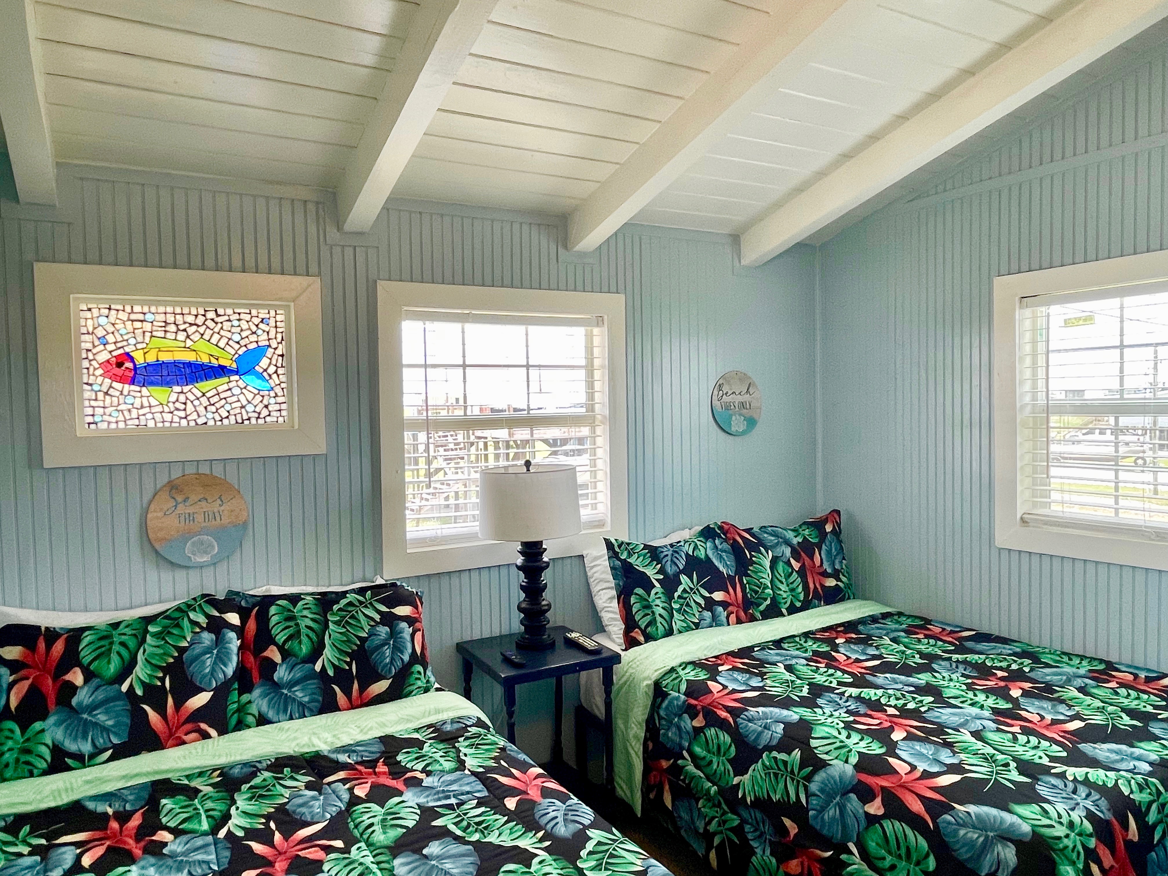 A brightly lit coastal bedroom with two beds, both featuring tropical-print bedding, light blue walls, and a white exposed beam ceiling.