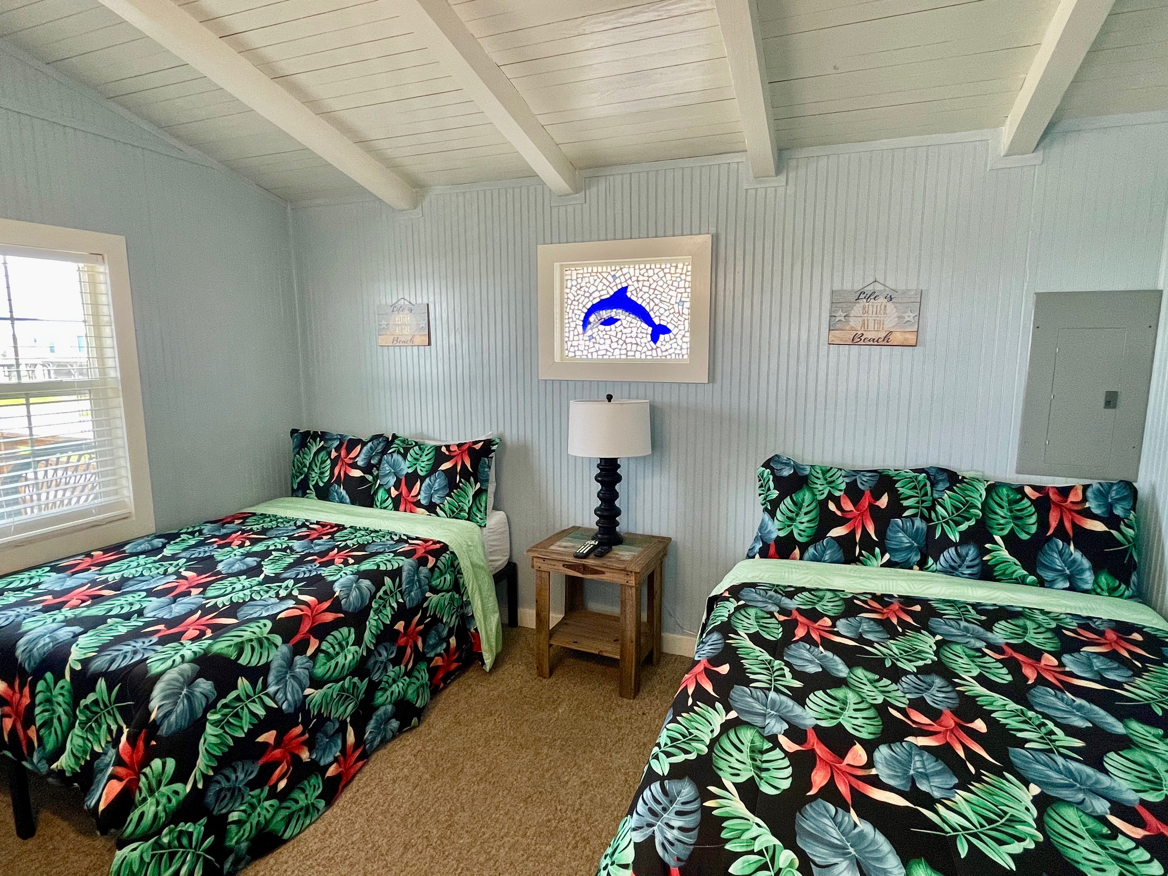A vibrant bedroom with two beds featuring tropical-print bedding, light blue walls, a sloped white ceiling with exposed beams, and dolphin stained glass window.