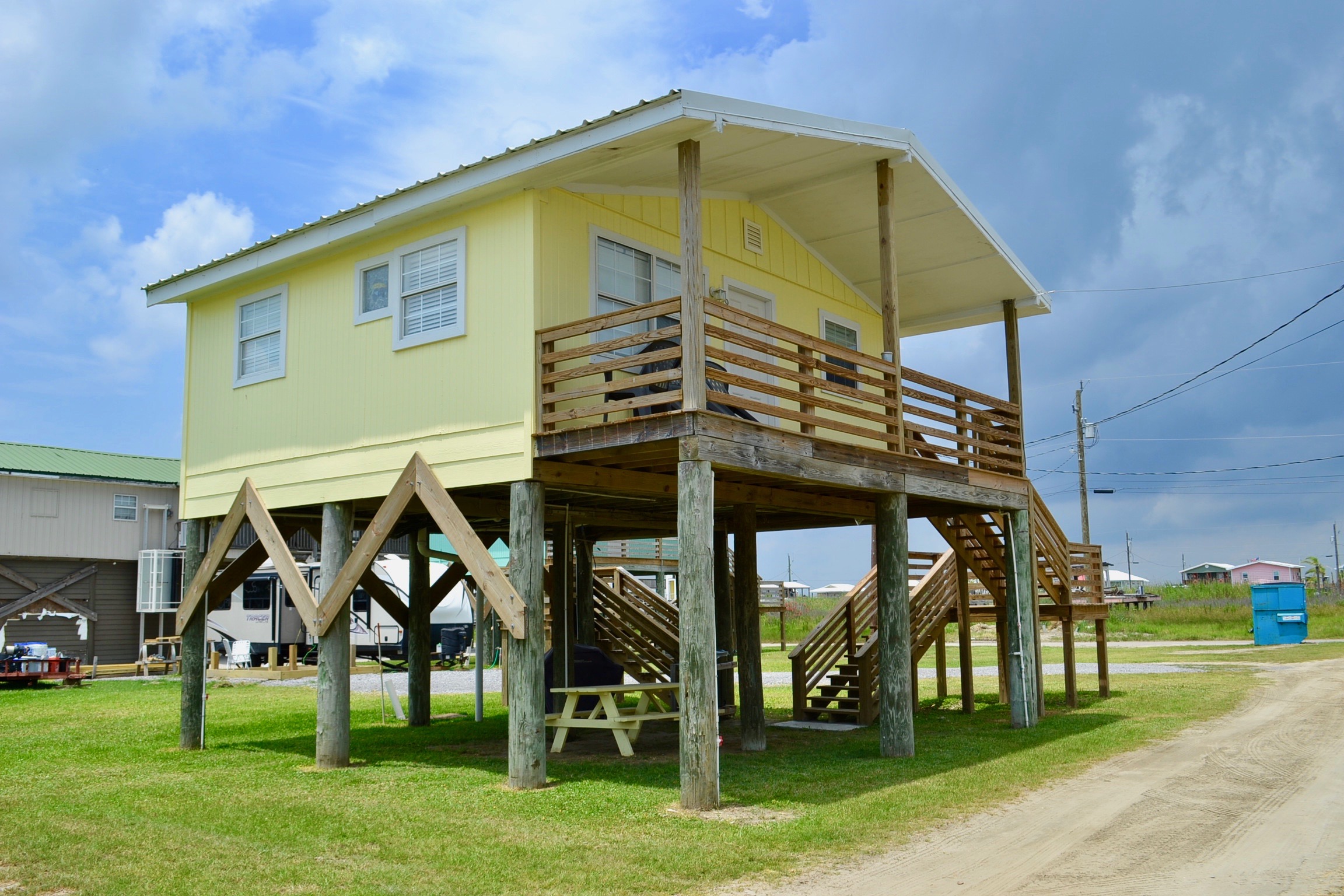 Bright yellow coastal stilt house with a covered deck, wooden pilings, and stairs, set on a grassy lot under a sunny, blue sky.
