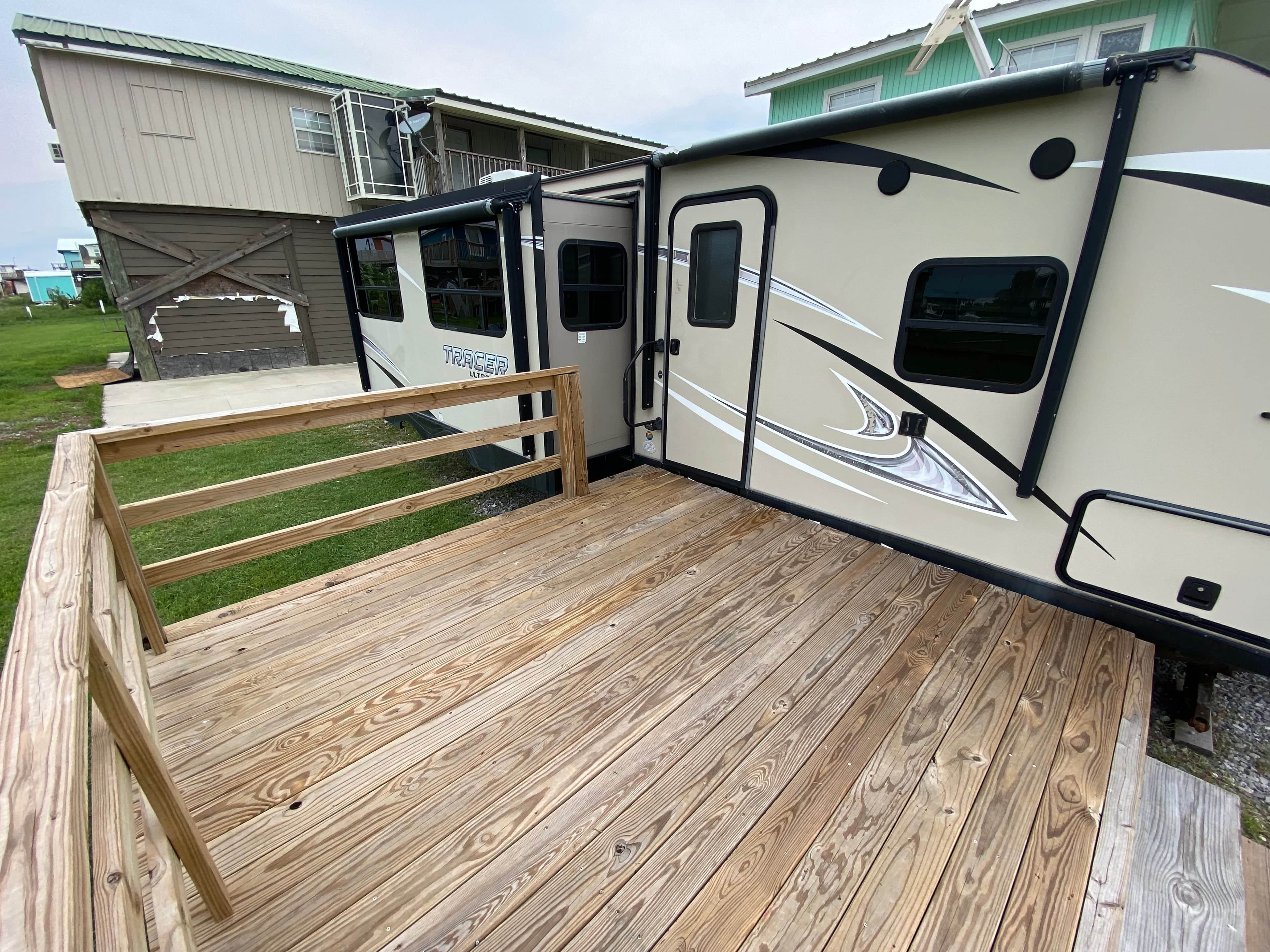 An elevated travel trailer with its slide-outs extended, parked next to a newly built wooden deck with a simple railing.