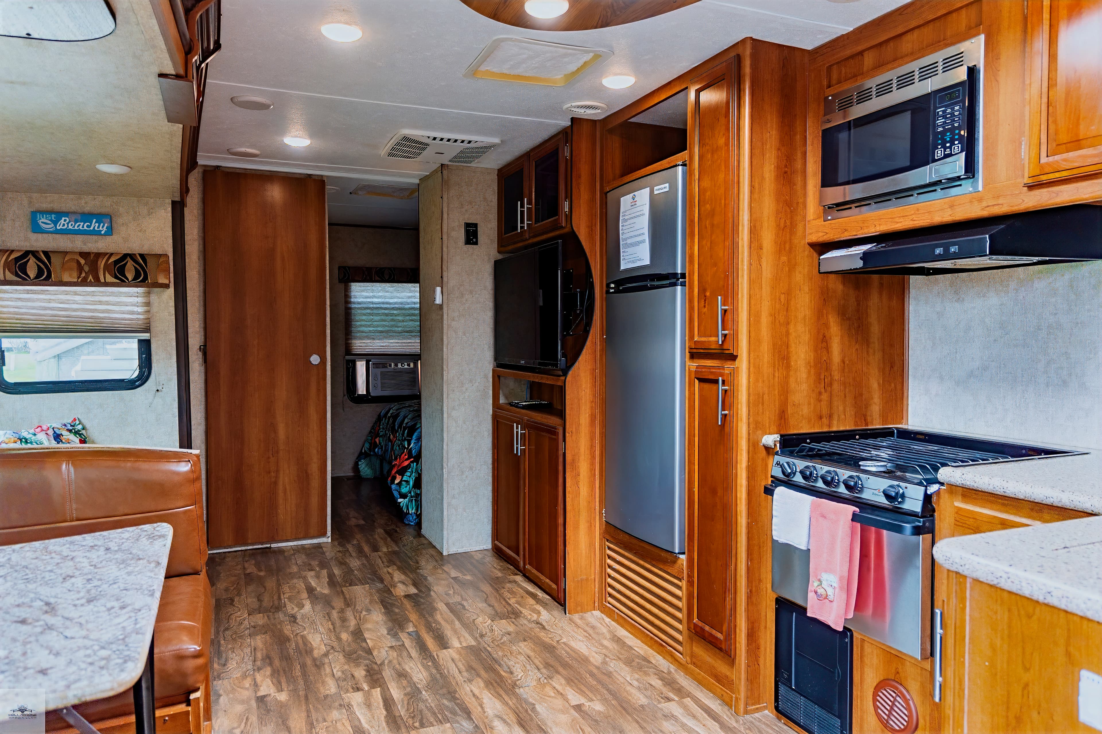 The galley-style kitchen of a camper features a stove, wood cabinets, a stainless steel fridge, and an open view toward a bedroom.
