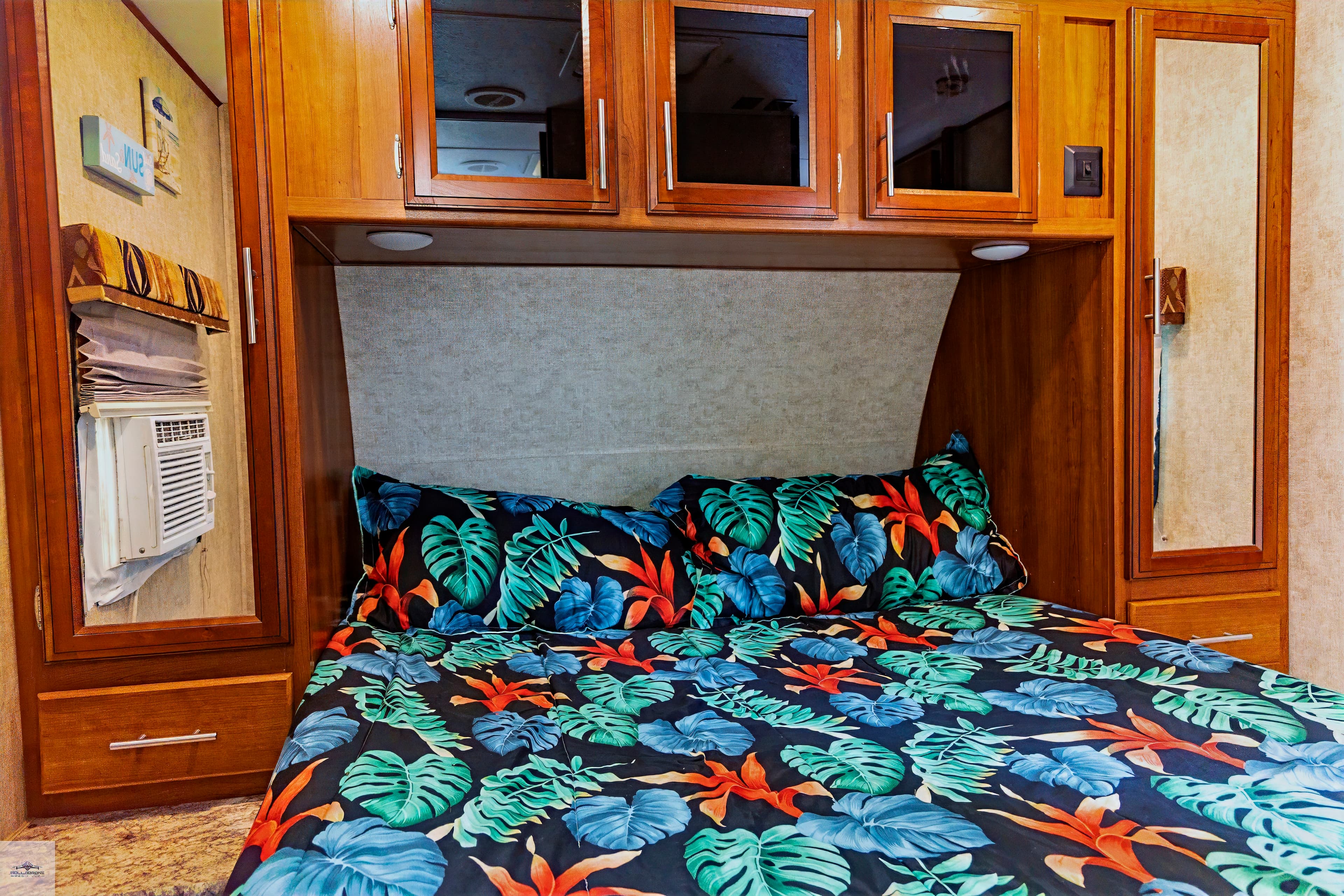 The compact bedroom of a camper featuring a bed with a tropical-print comforter, wood cabinetry overhead, and mirrored closets on each side.