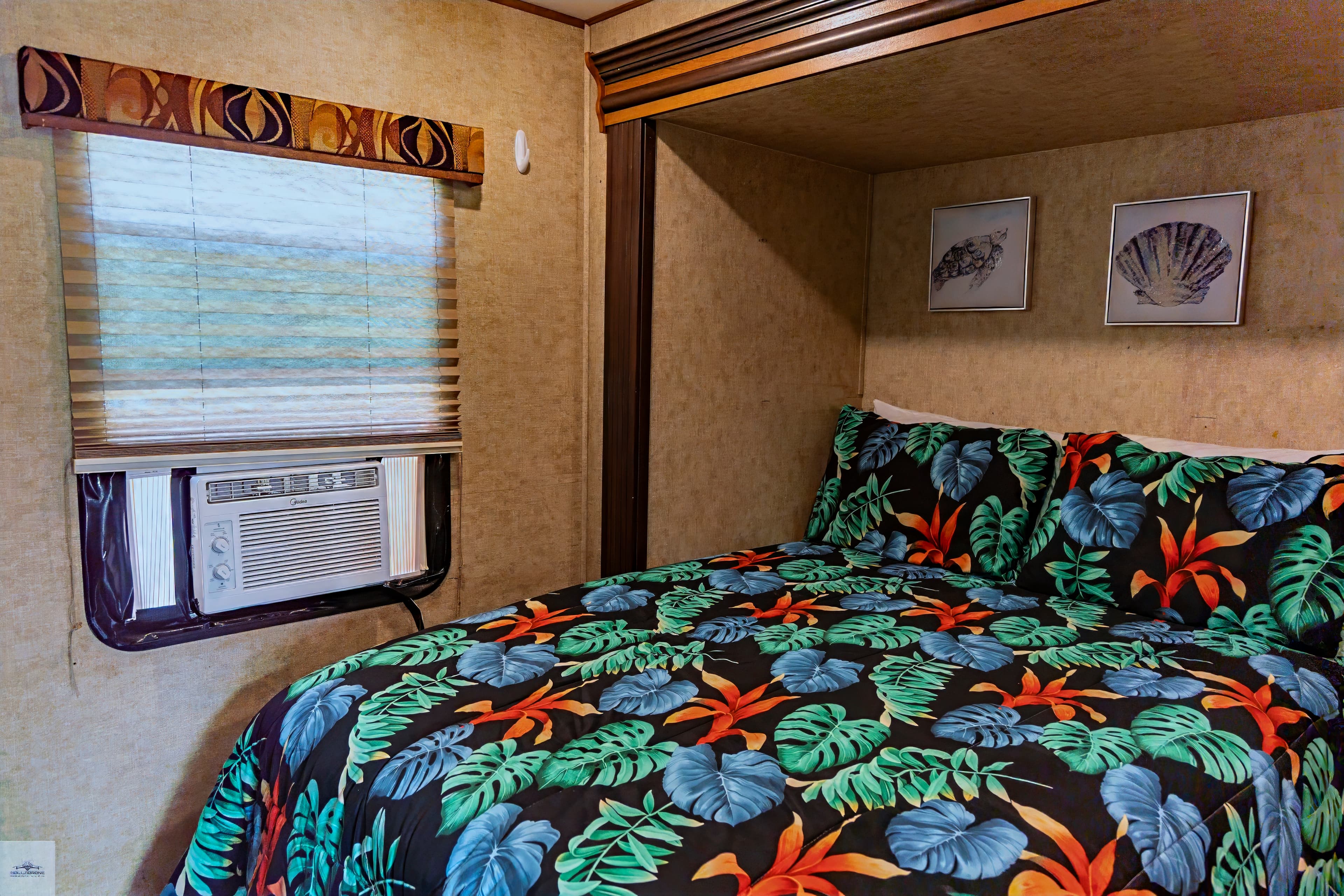 A compact camper bedroom featuring a bed with a tropical-print comforter nestled in a niche, a window AC unit, and shell wall art.