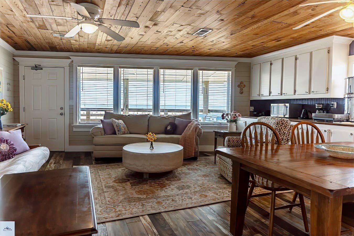 An open-concept living space with a light sofa near a bank of windows, a wooden ceiling, a dining area, and a full kitchen visible.