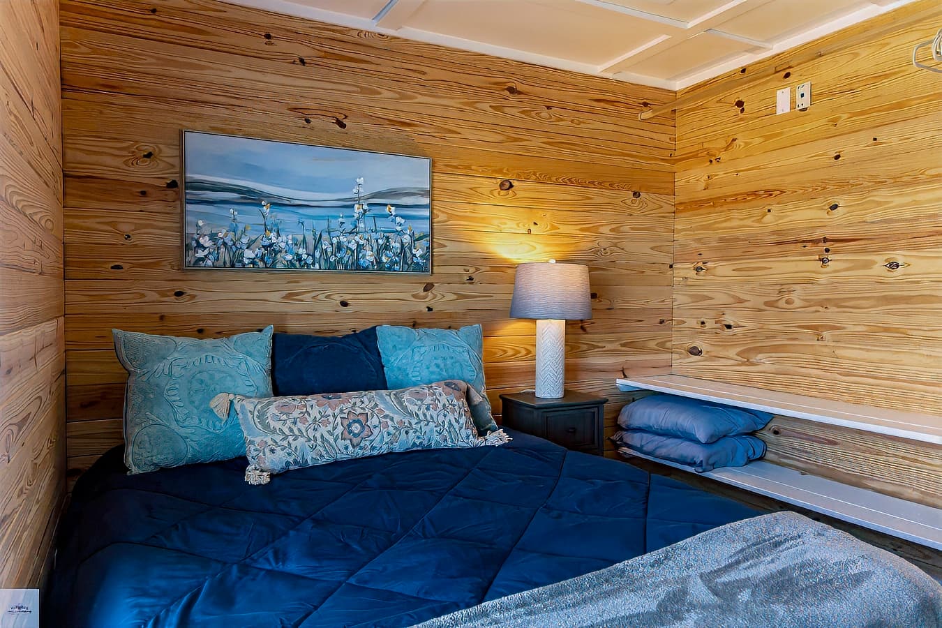 A rustic yet cozy bedroom featuring knotty pine walls, a deep blue comforter, coastal artwork, and built-in shelves.