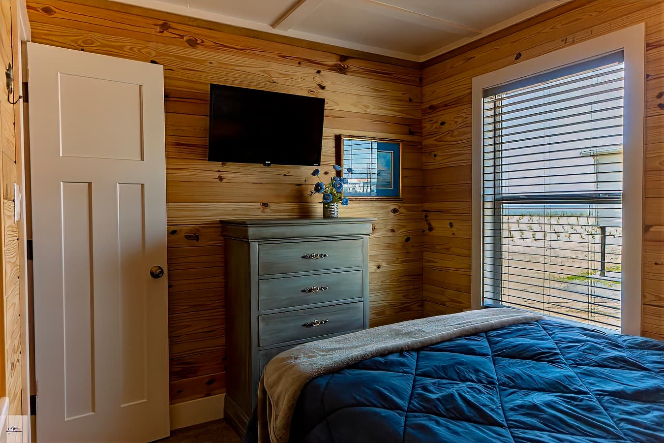 A bedroom with knotty pine wood walls, a blue comforter, a gray dresser with a TV mounted above it, and a side window with blinds.