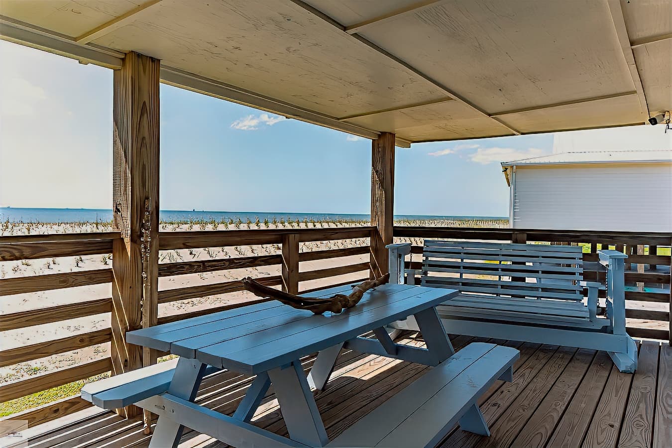 A covered deck with a light blue picnic table and a matching glider, offering a beautiful gulf view over a wooden railing.
