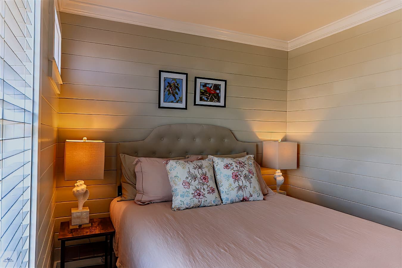 A serene bedroom with a tufted headboard, light pink bedding, matching lamps, and two framed prints on the light, shiplap-style walls.