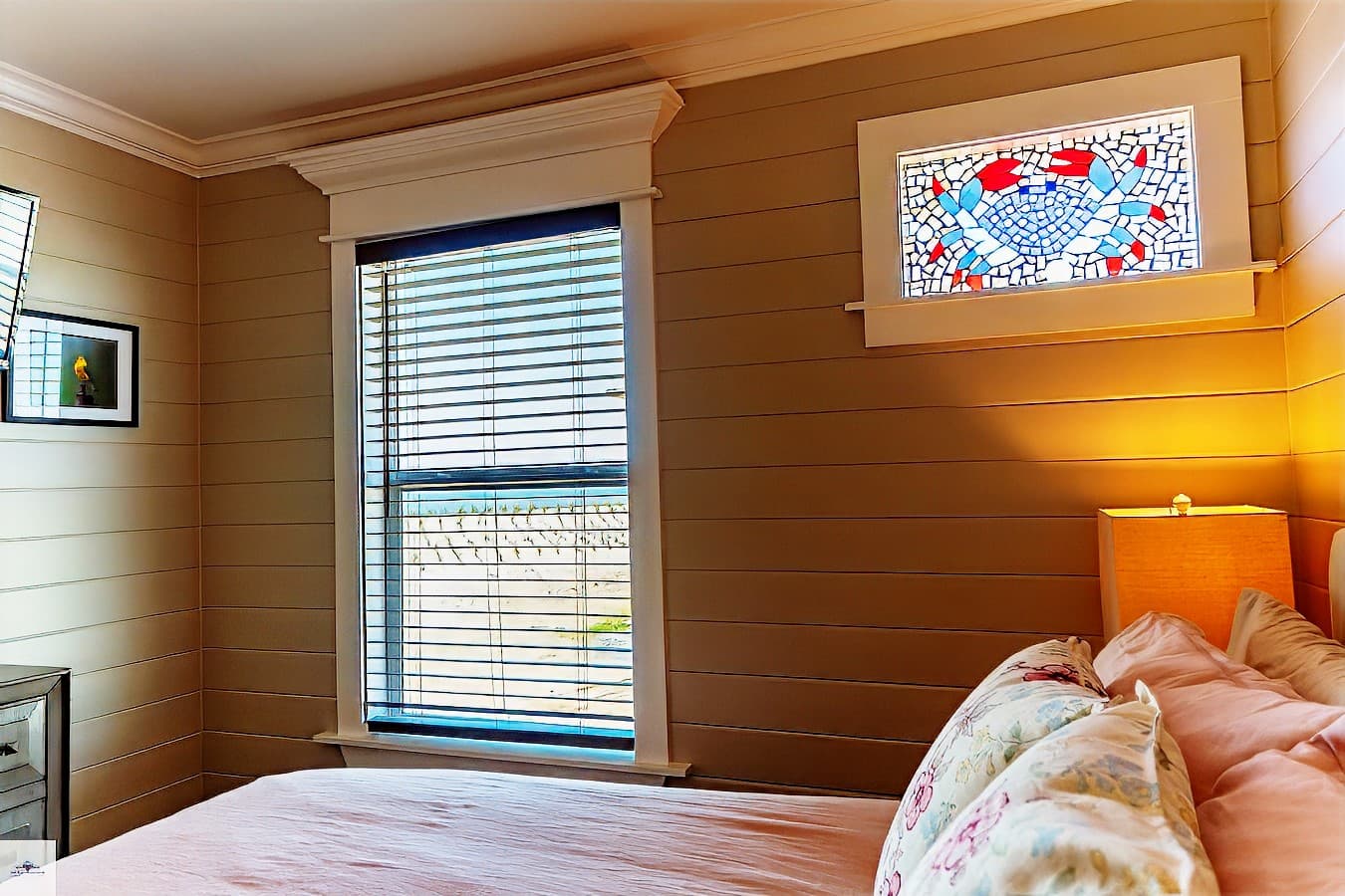 A bedroom corner showing a bed with pink bedding, a window with blinds, and a unique piece of crab-themed stained glass window.