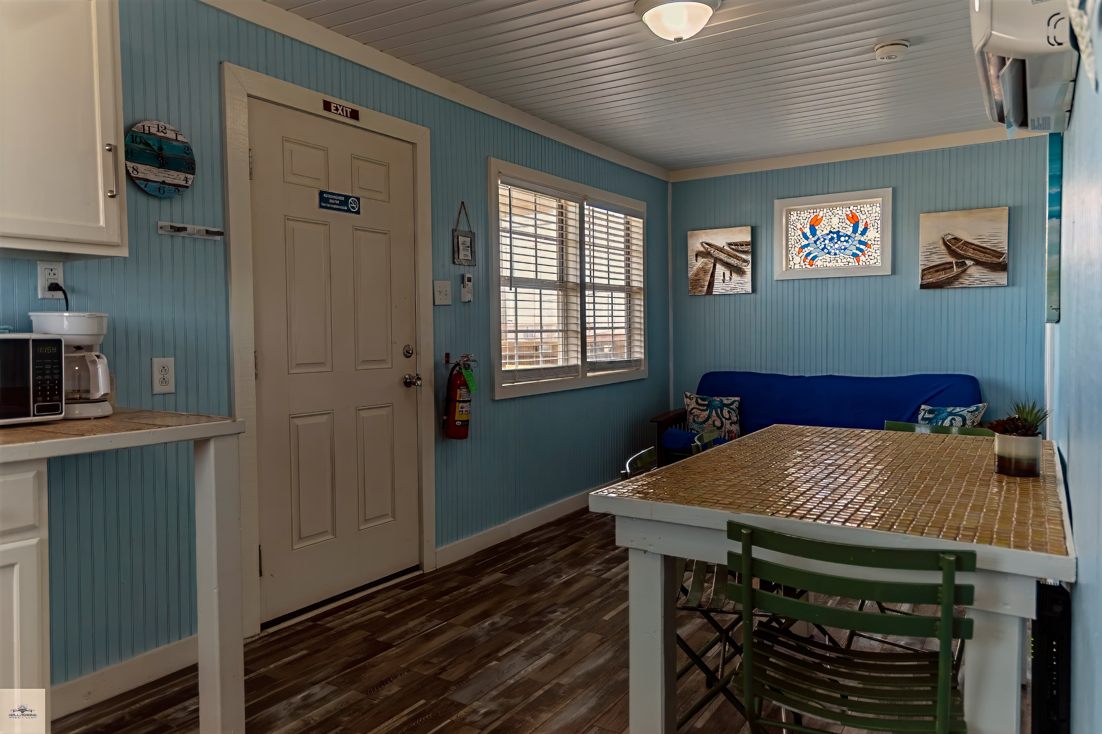 A small studio unit with light blue walls, a dining table, a blue futon in the background, and a compact kitchenette on the left.