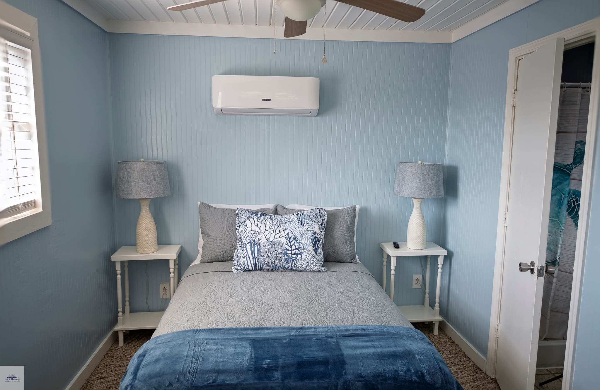 A bedroom with light blue walls, a bed with grey and blue bedspread, nightstands with lamps on either side of the bed, and an AC unit above the bed.