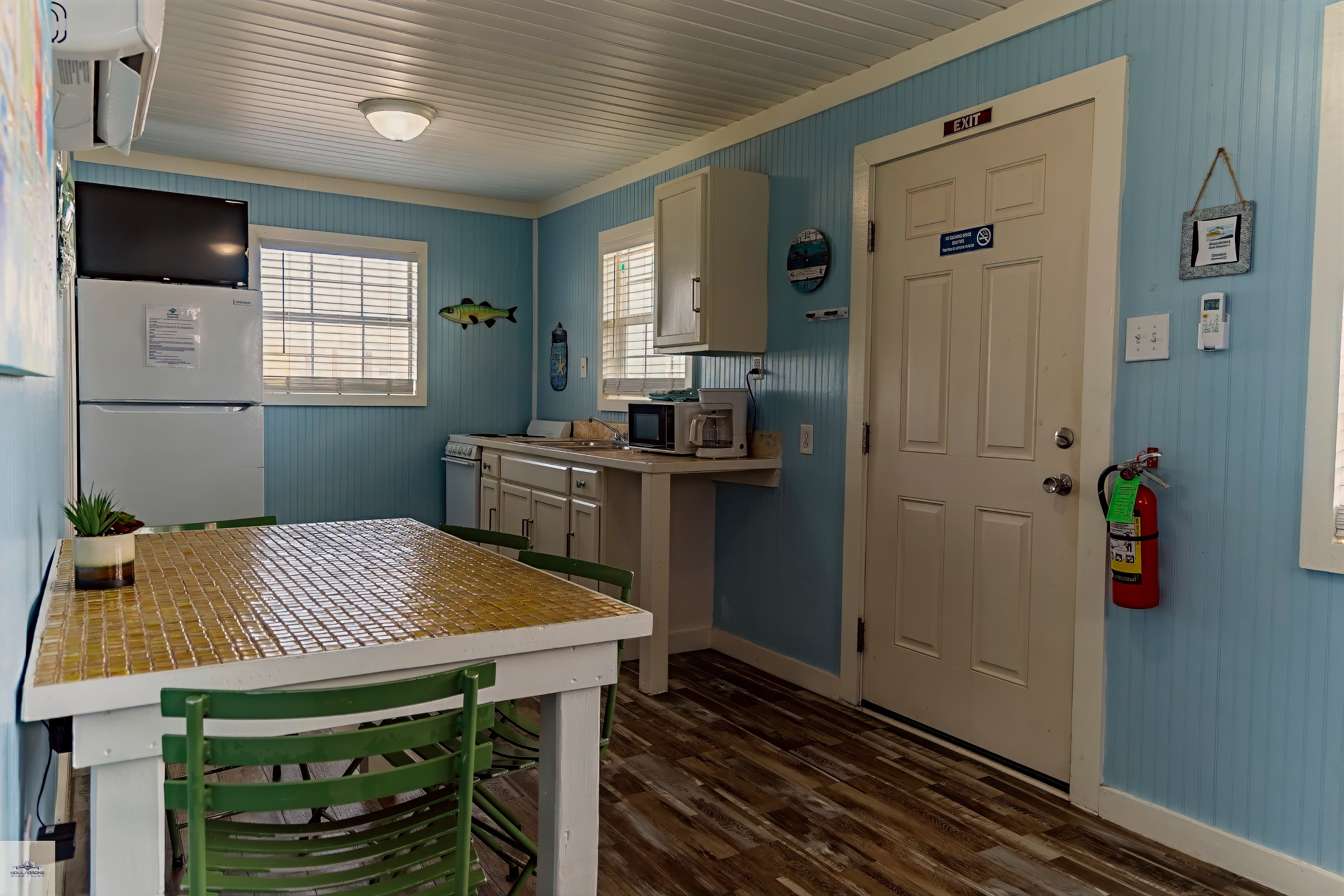 Cozy kitchen with light blue beadboard walls and a white dining table with green chairs in a beach-themed cottage.