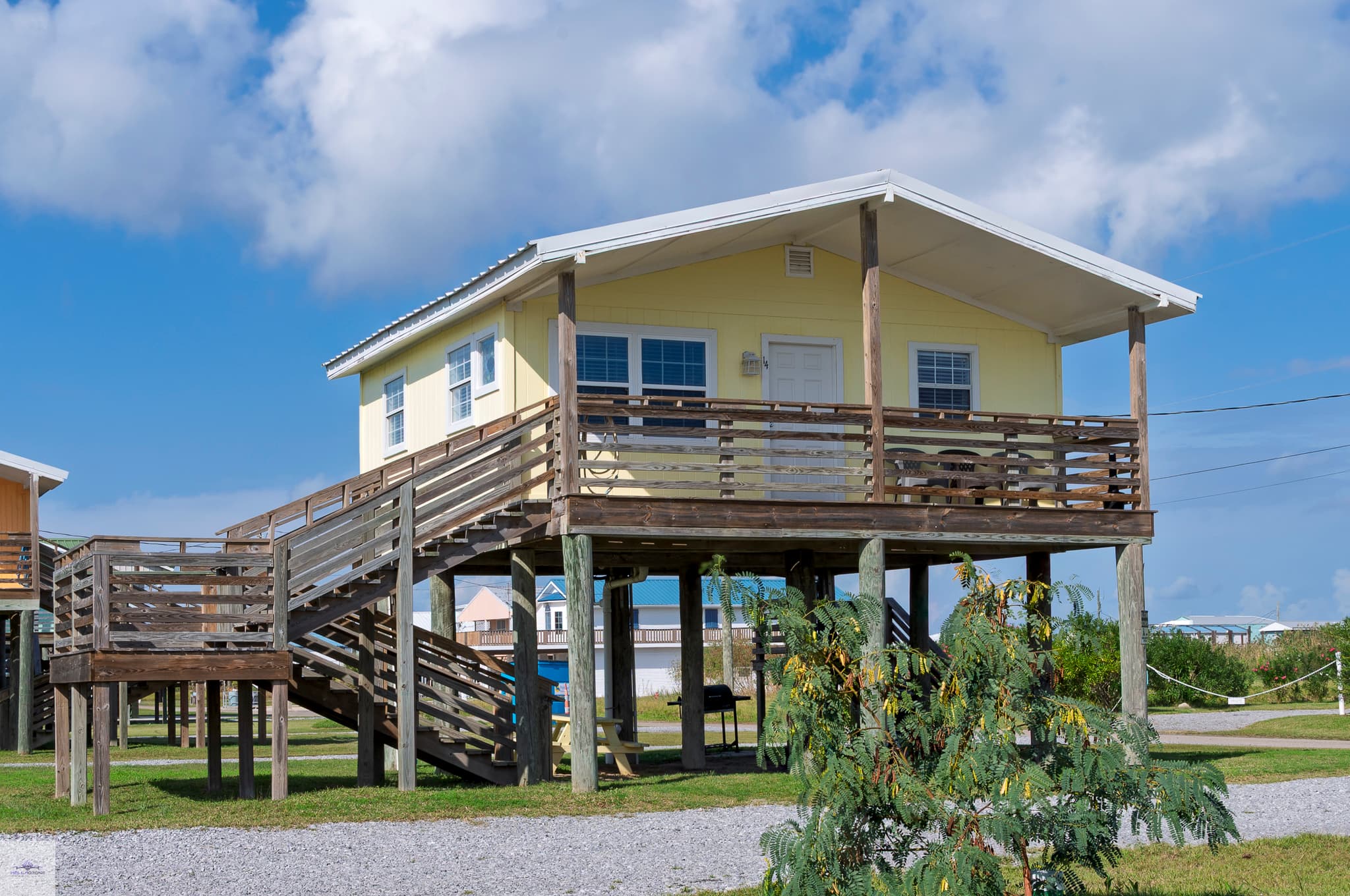 Bright yellow coastal stilt house with a covered deck, wooden pilings, and stairs, set on a grassy lot under a sunny, blue sky.