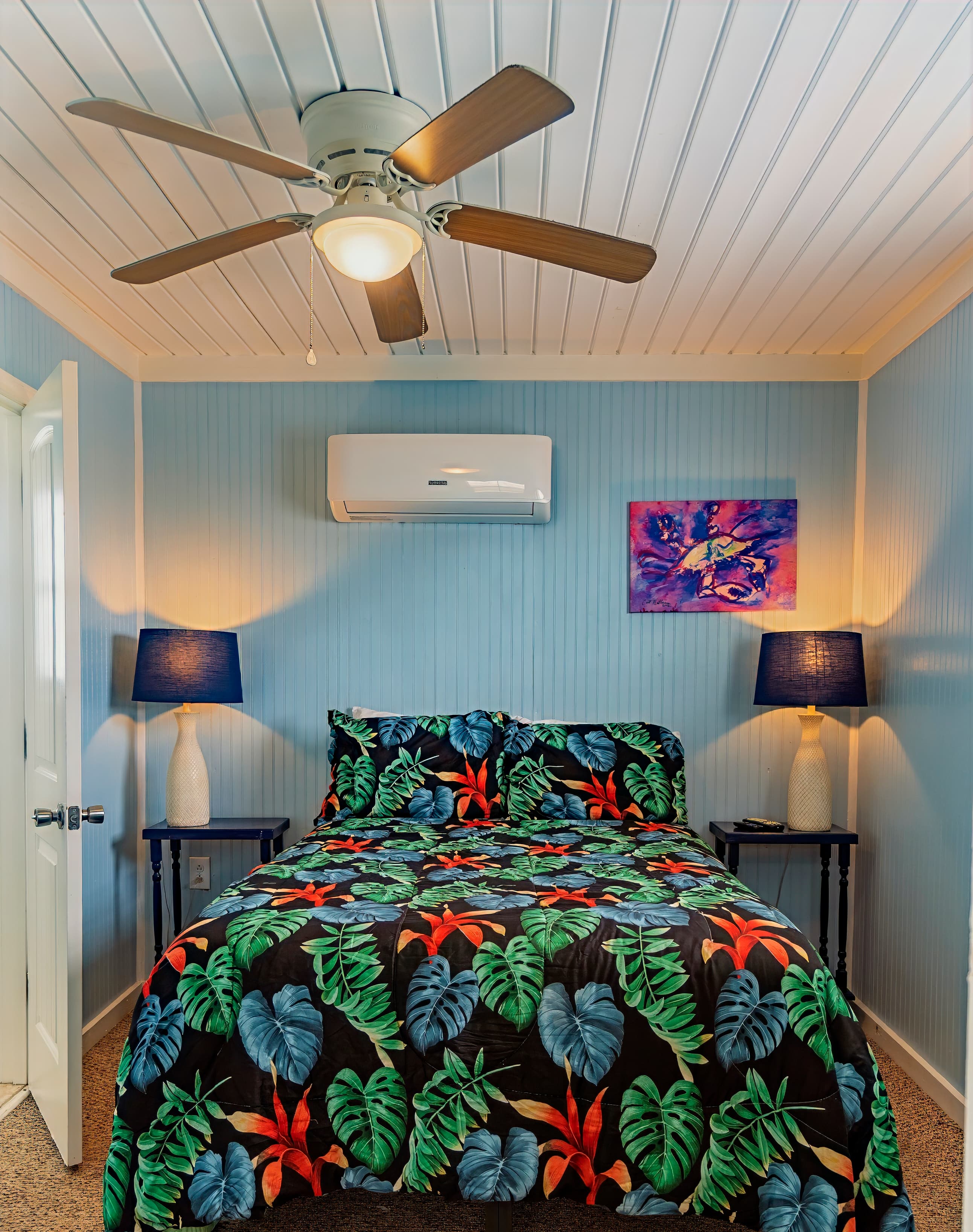 Tropical-themed bedroom with sky blue walls, a colorful duvet, white ceiling fan, and two navy blue lamps on flanking nightstands.