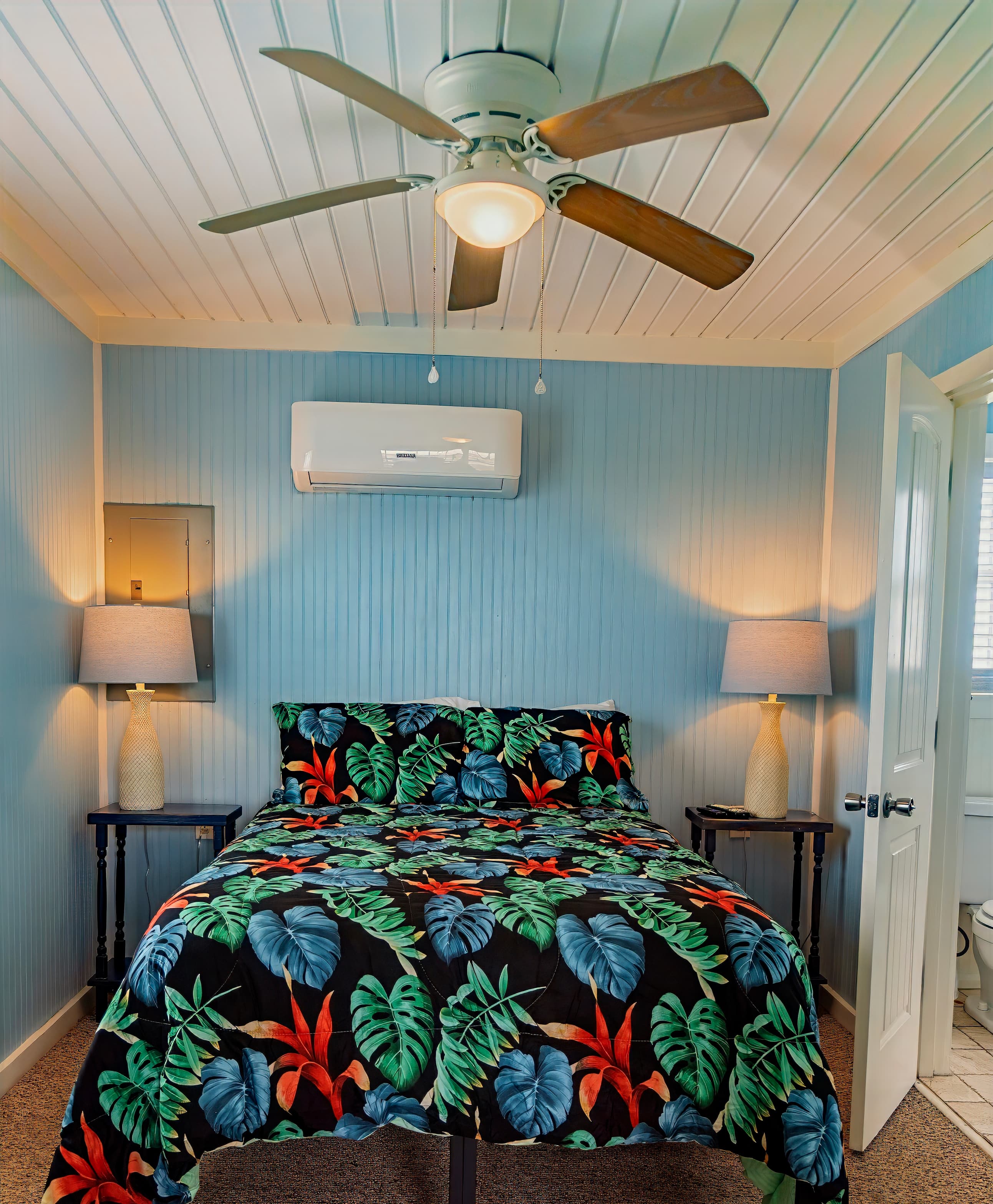 Bright beach cottage bedroom with sky blue walls, a tropical leaf-print bedspread, ceiling fan, and access to an ensuite bathroom.