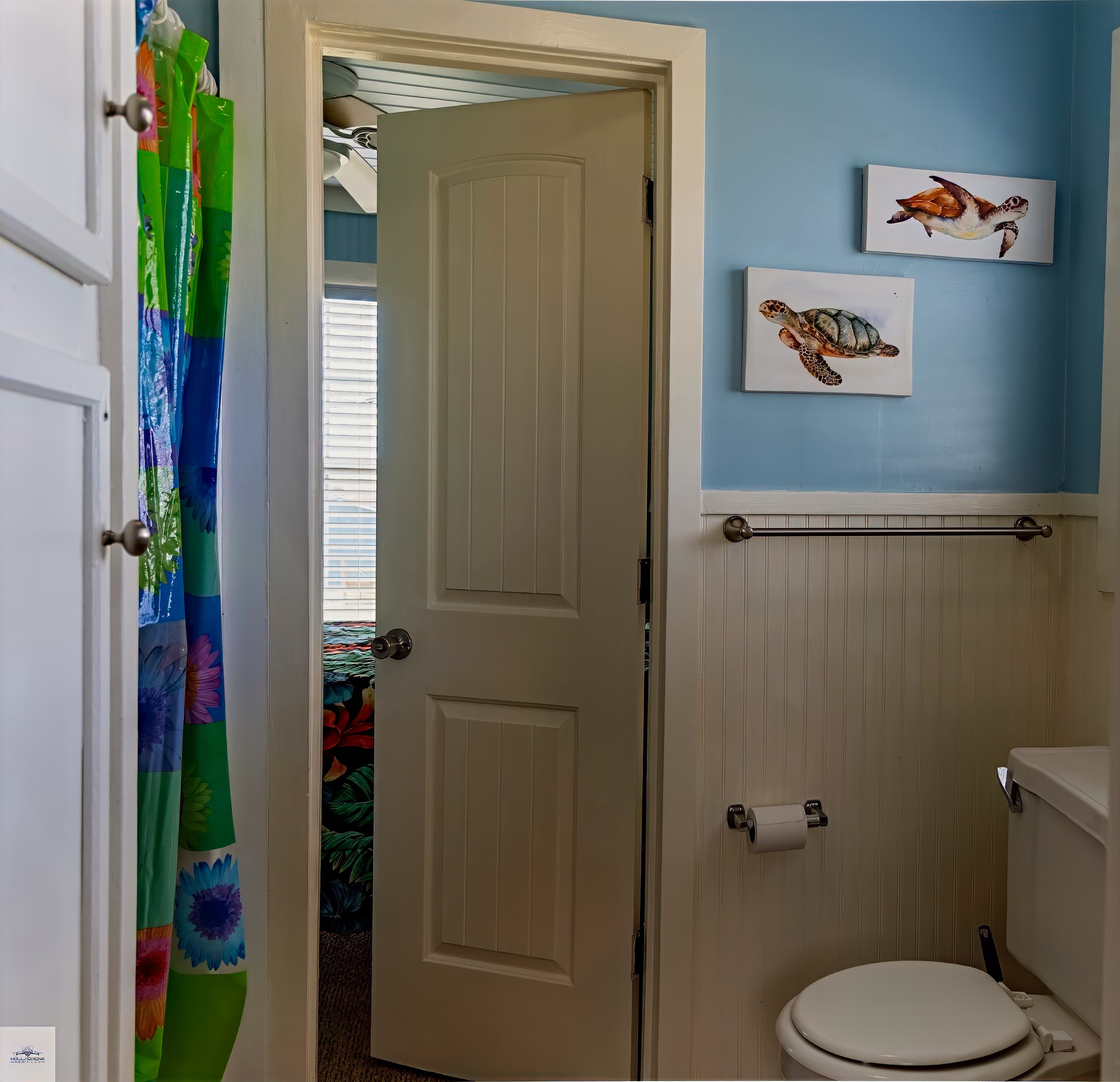 Beach-themed bathroom with light blue and white beadboard walls, a toilet, and sea turtle art. An open doorway leads to a bedroom.