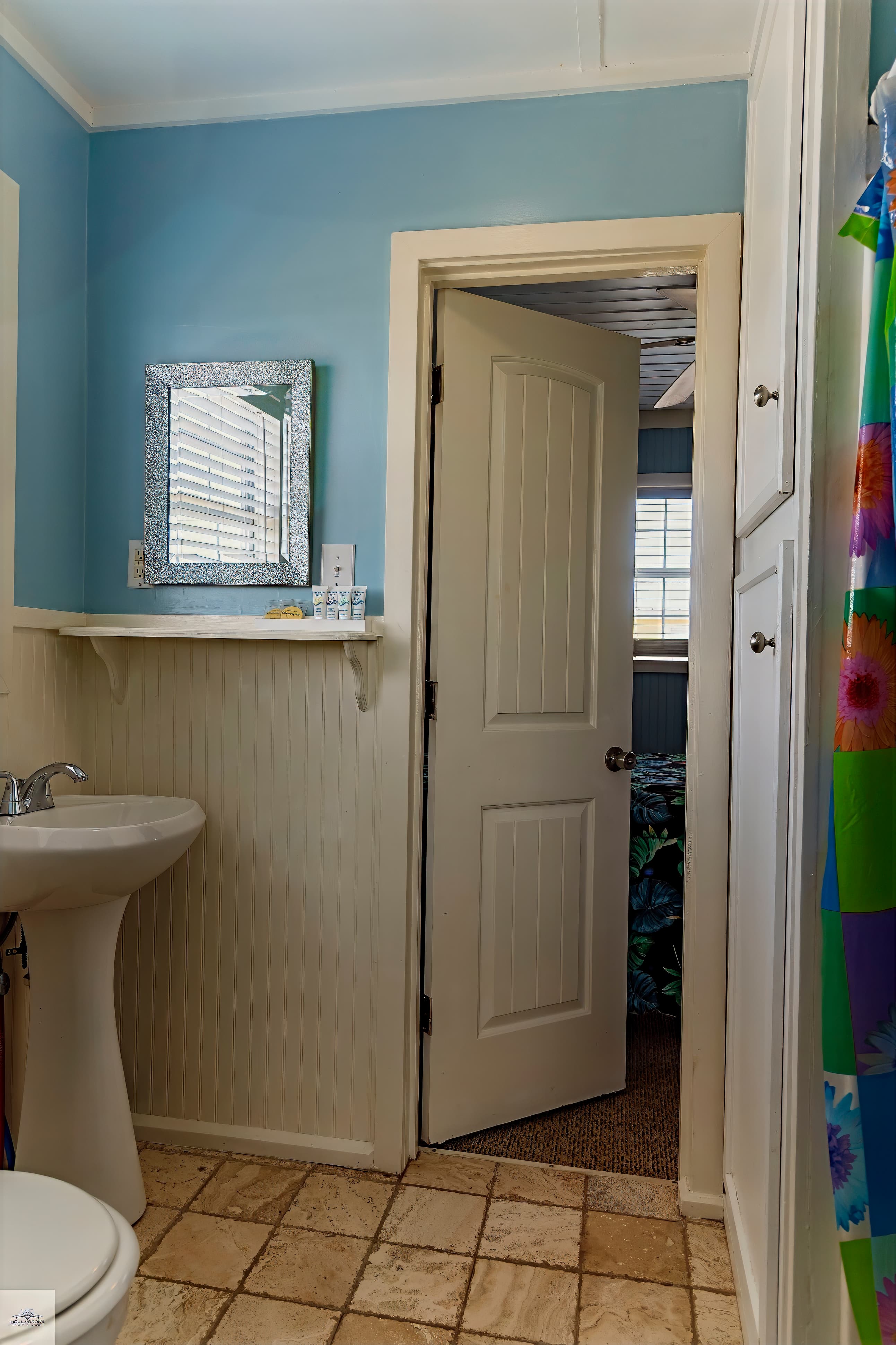 Sky blue bathroom with a pedestal sink, white beadboard wainscoting, and an open door leading into a bedroom with a window.