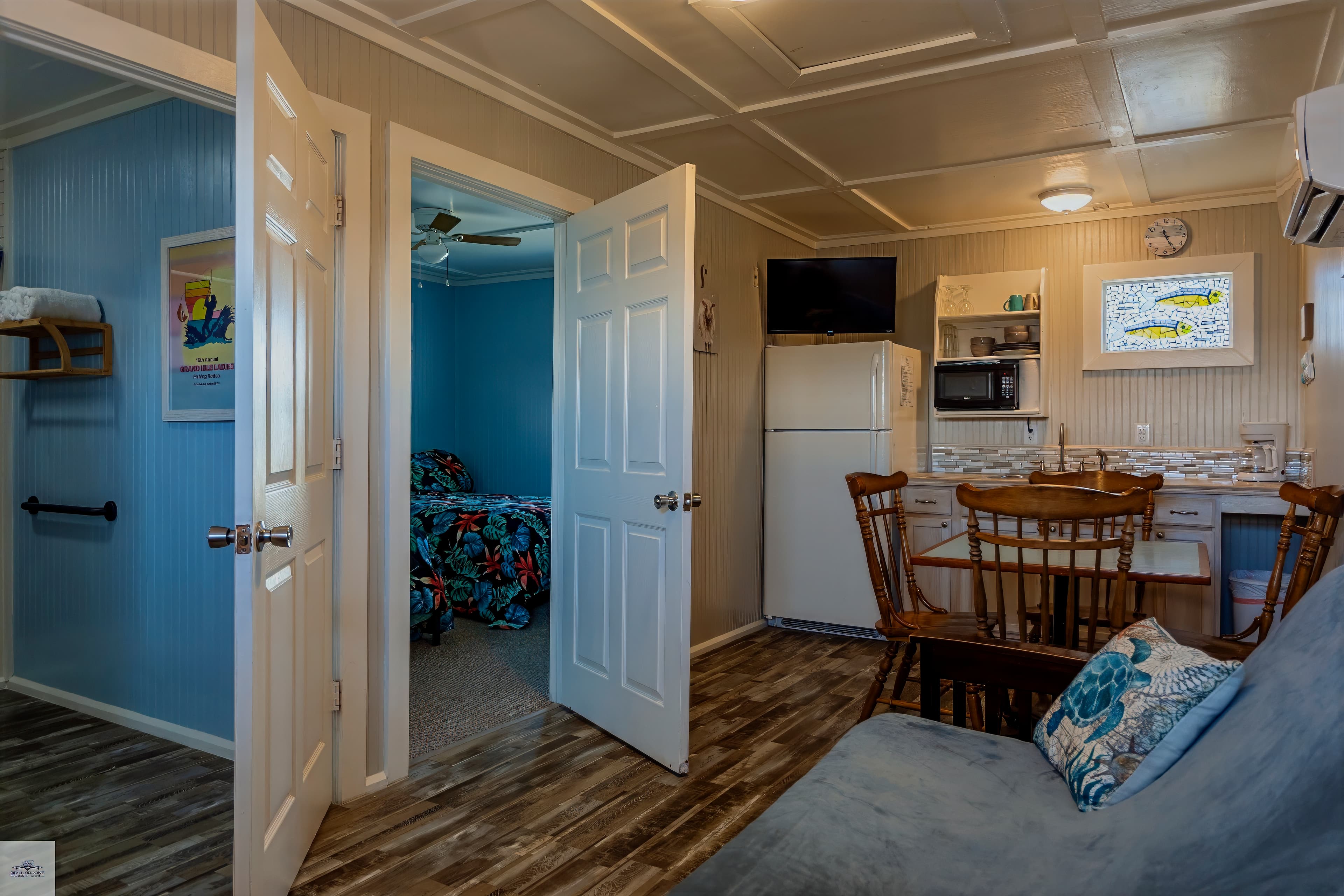 Living area with a kitchenette, dining table, and futon, showing an open doorway into a bedroom with a floral bedspread.