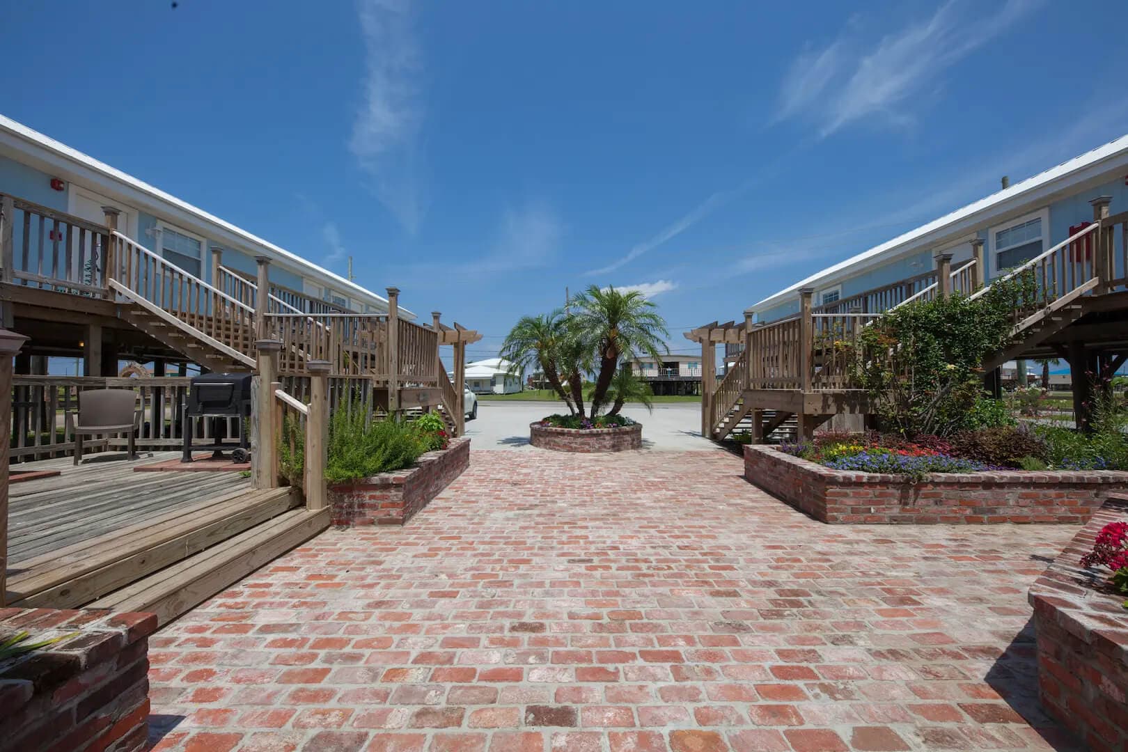 A brick pathway leads to a central palm tree, bordered by wooden decks and clear blue sky.