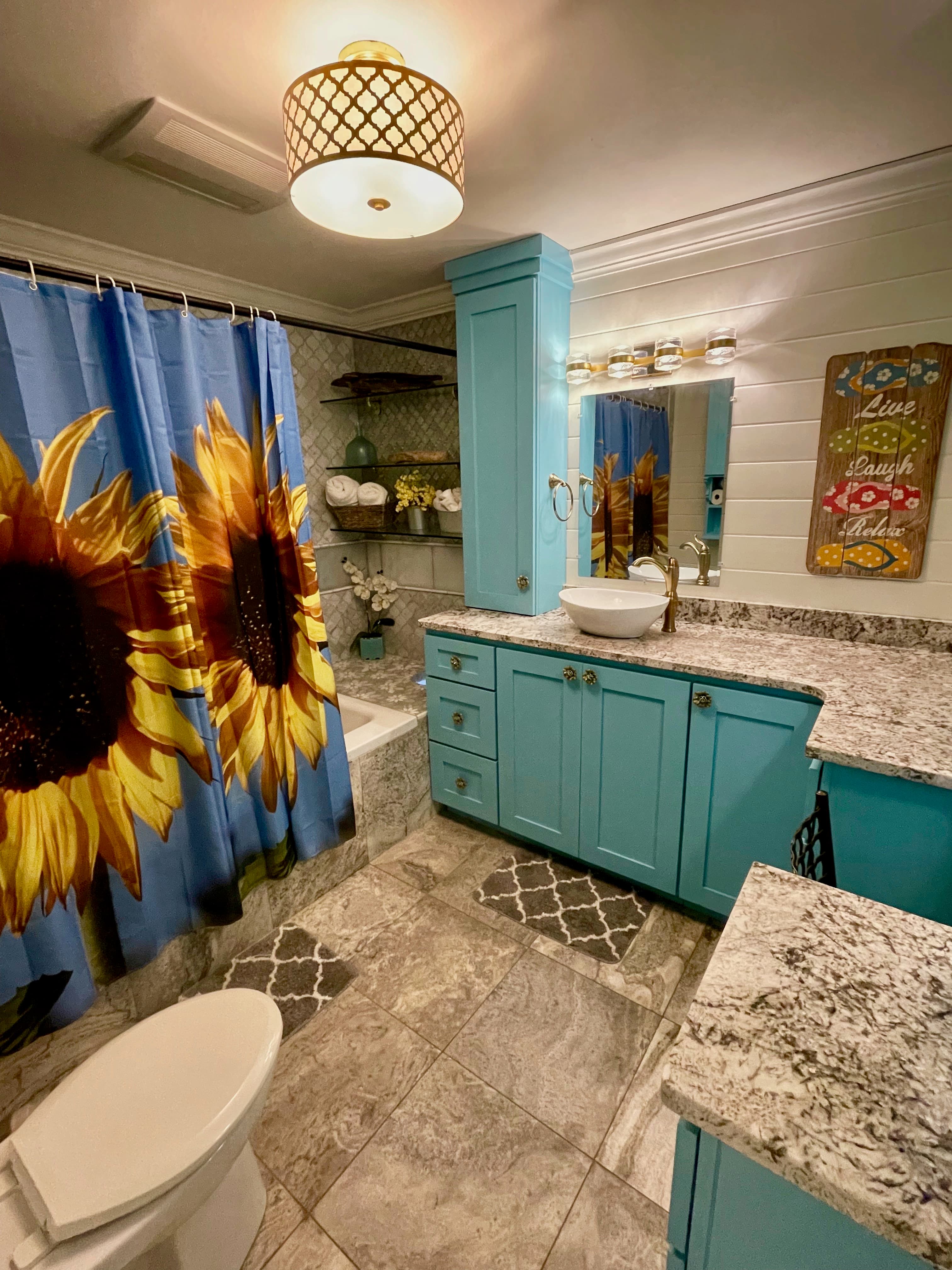 A vibrant bathroom featuring bold turquoise cabinetry and granite counters, a sunflower-print shower curtain, and a vessel sink.