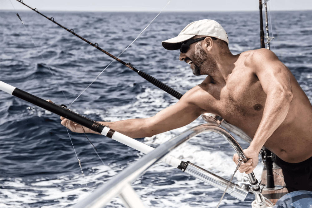 A shirtless man with sunglasses joyfully reels in a fishing line on a boat in the ocean.