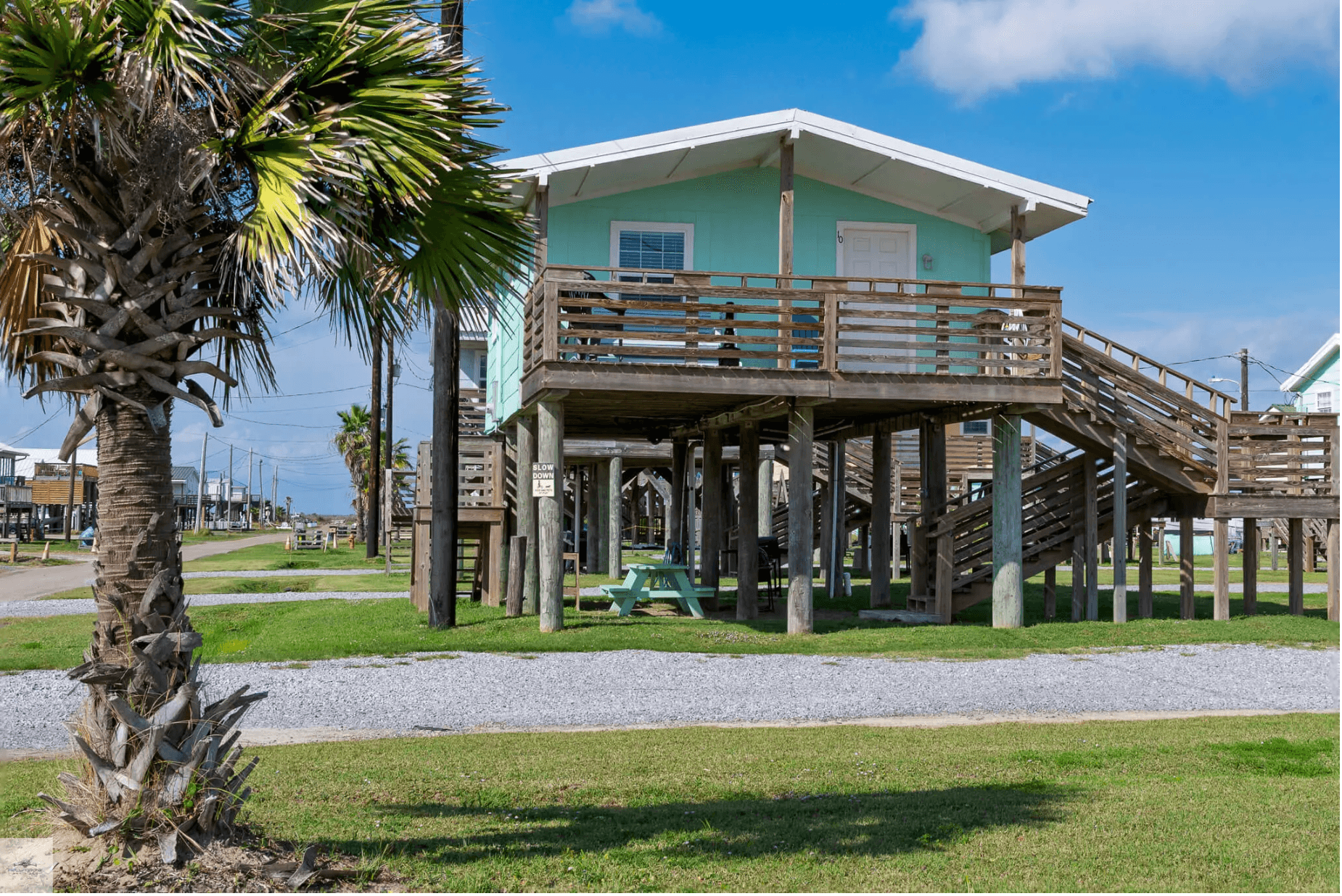 A light blue house on stilts with a wooden deck, surrounded by palm trees and grass under a clear blue sky.