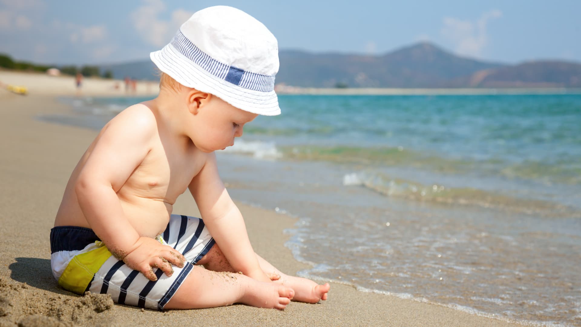 A baby sits on the beach, playing in the sand with the ocean in the background.
