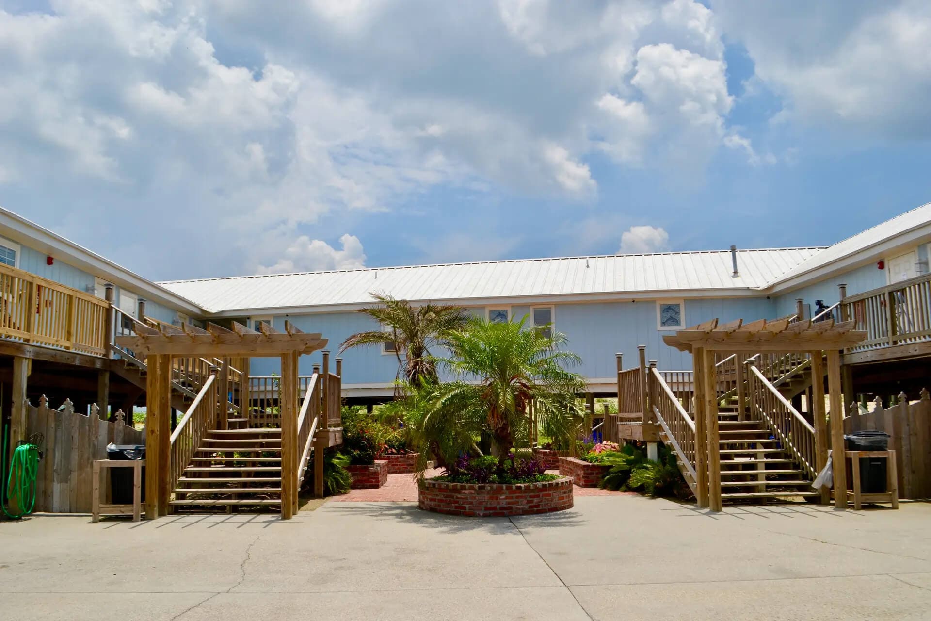 A courtyard with wooden staircases leading to a building, featuring a central planter with tropical plants under a cloudy sky.