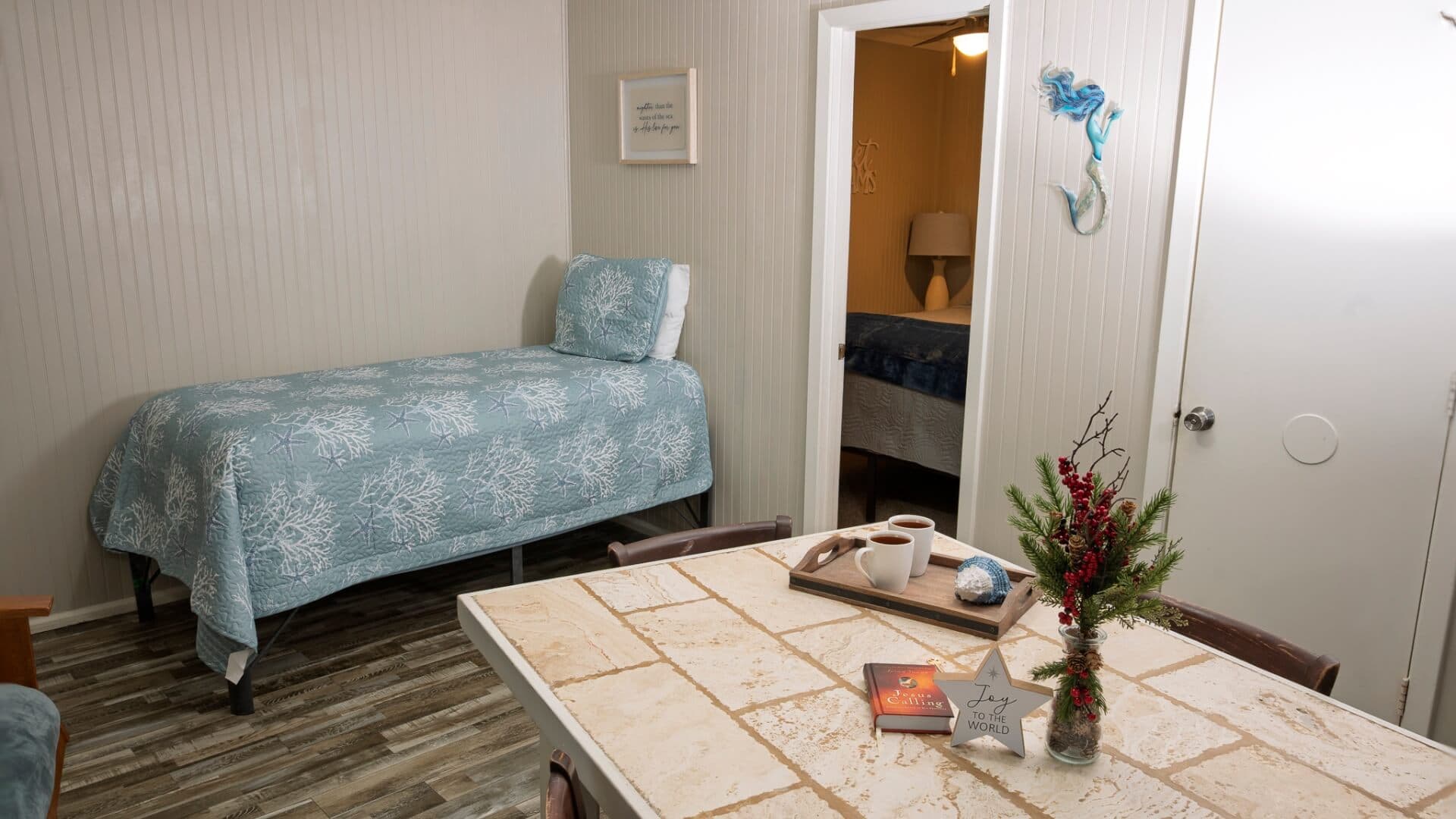 A cozy room featuring a single bed with a light blue coverlet, a dining table, and an open doorway to another bedroom.