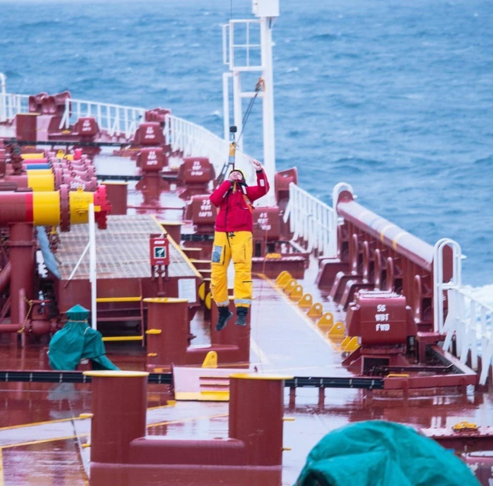 A person in a red jacket is being lifted by a harness on a ship's deck in rough weather.