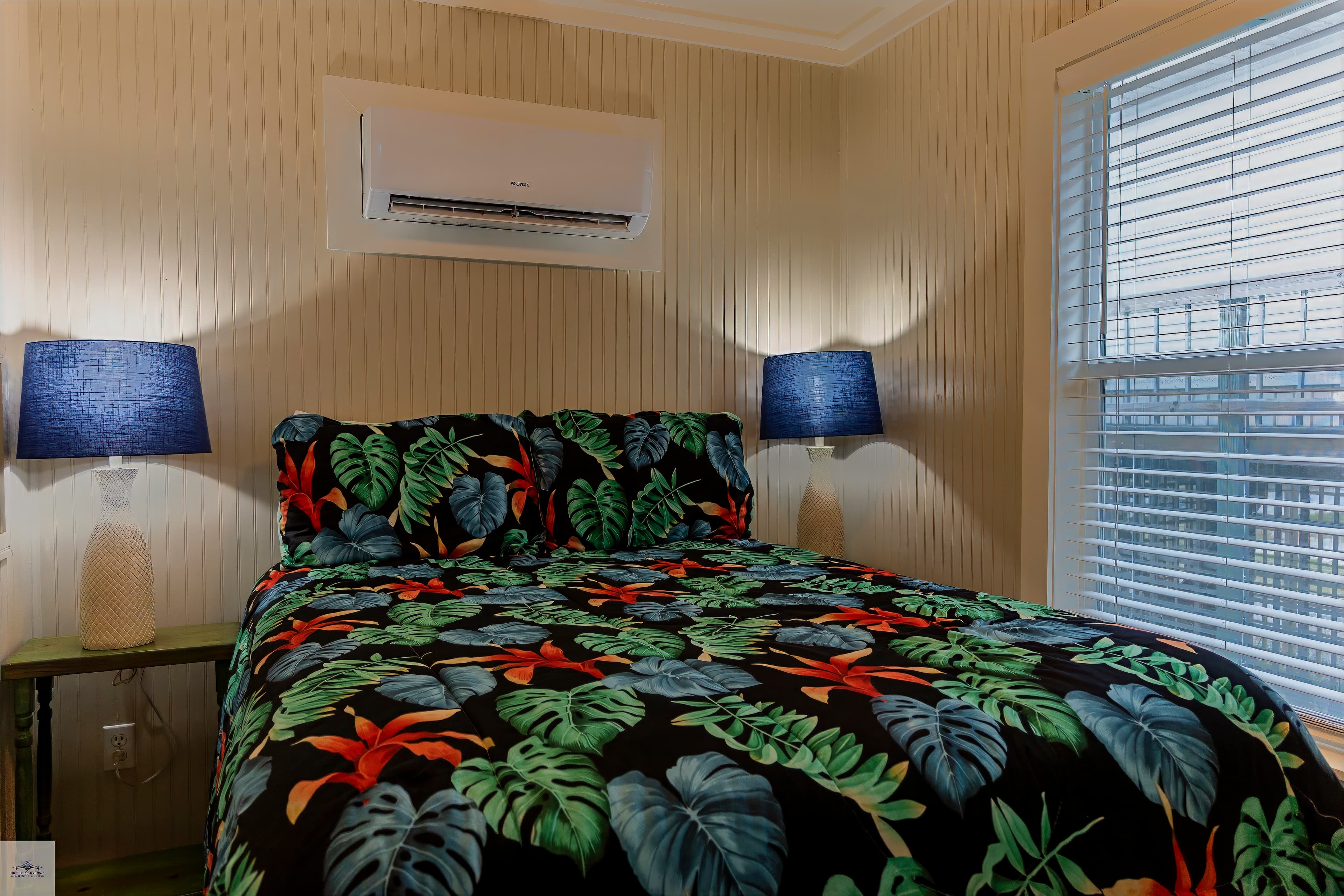 A cozy bedroom featuring a bed with tropical-themed bedding, two blue lamps, and an air conditioning unit.