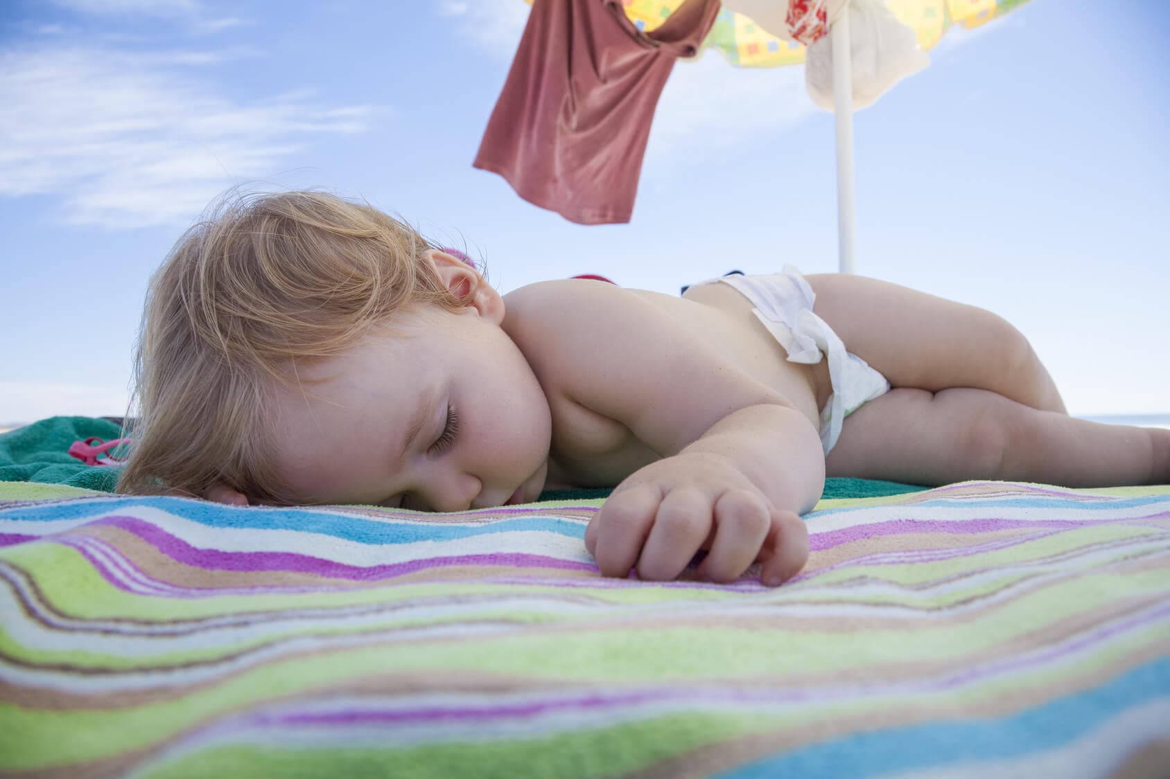 A sleeping child lays on a colorful beach towel under a bright blue sky.