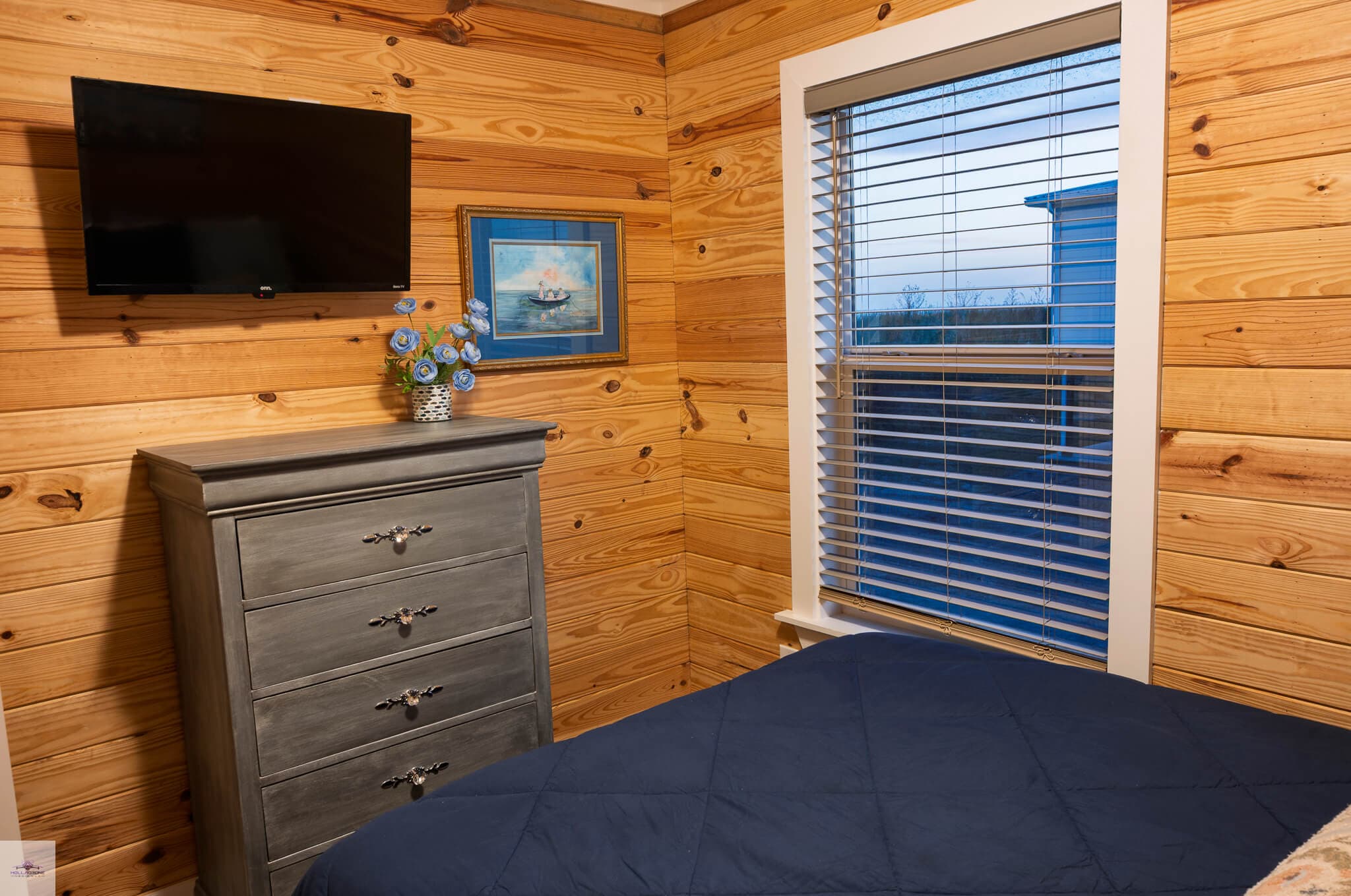 A cozy bedroom featuring a gray dresser, a flat-screen TV, a flower vase on a shelf, and wooden walls.