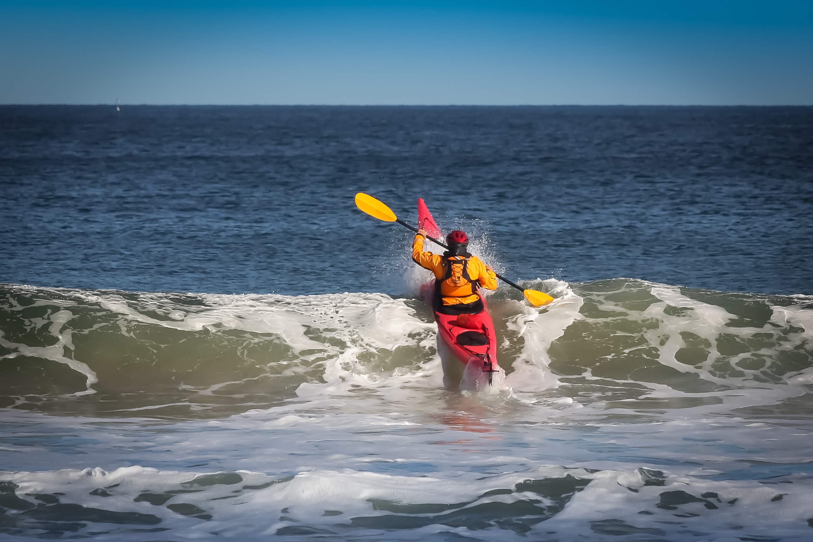 A kayaker navigates through ocean waves under a clear blue sky.