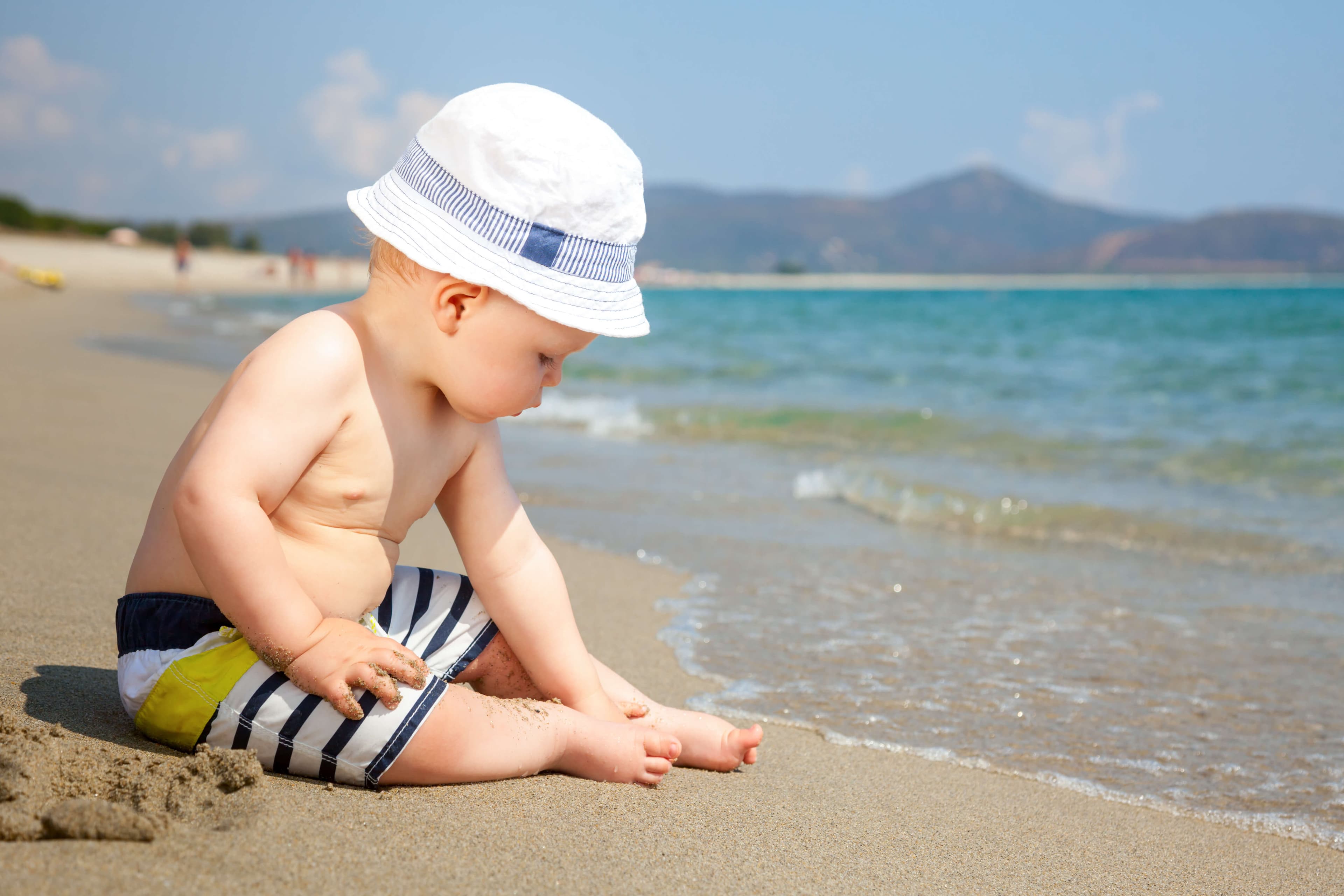 A young child sits on the sandy beach, playing with sand near the gentle waves of the ocean.