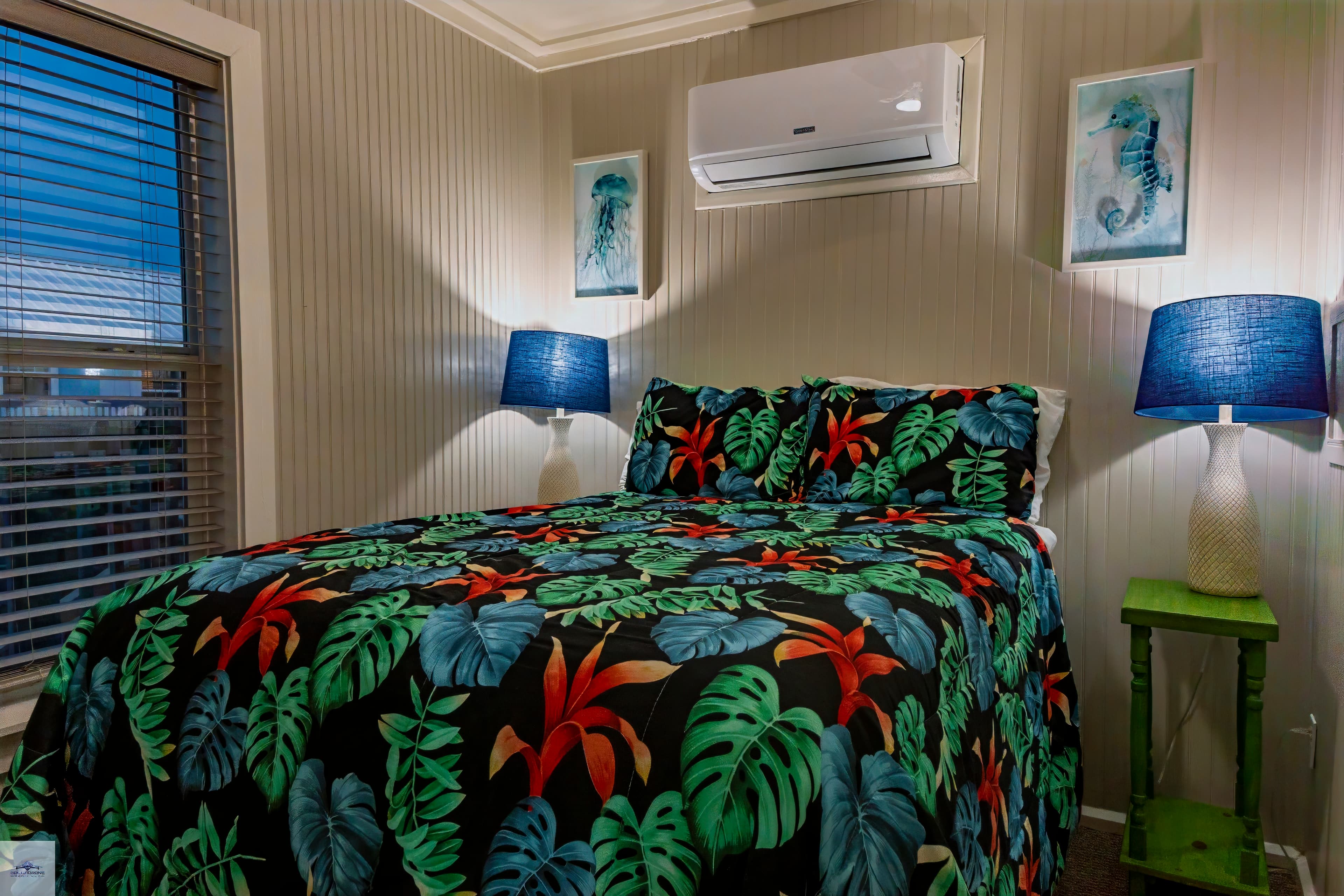 A close-up of a bedroom with a bed featuring a tropical floral bedspread, flanked by two matching lamps, and an AC unit above.
