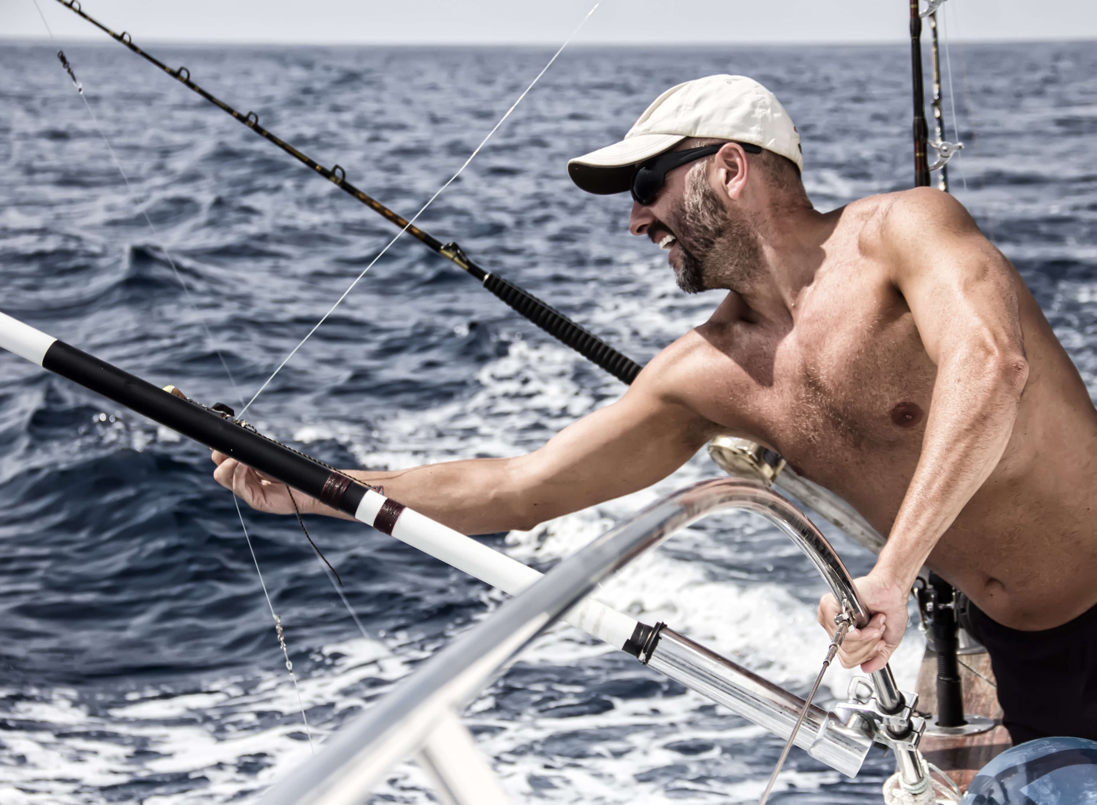 A shirtless man wearing a cap and sunglasses leans over the edge of a boat, holding a fishing rod on a sunny day at sea.
