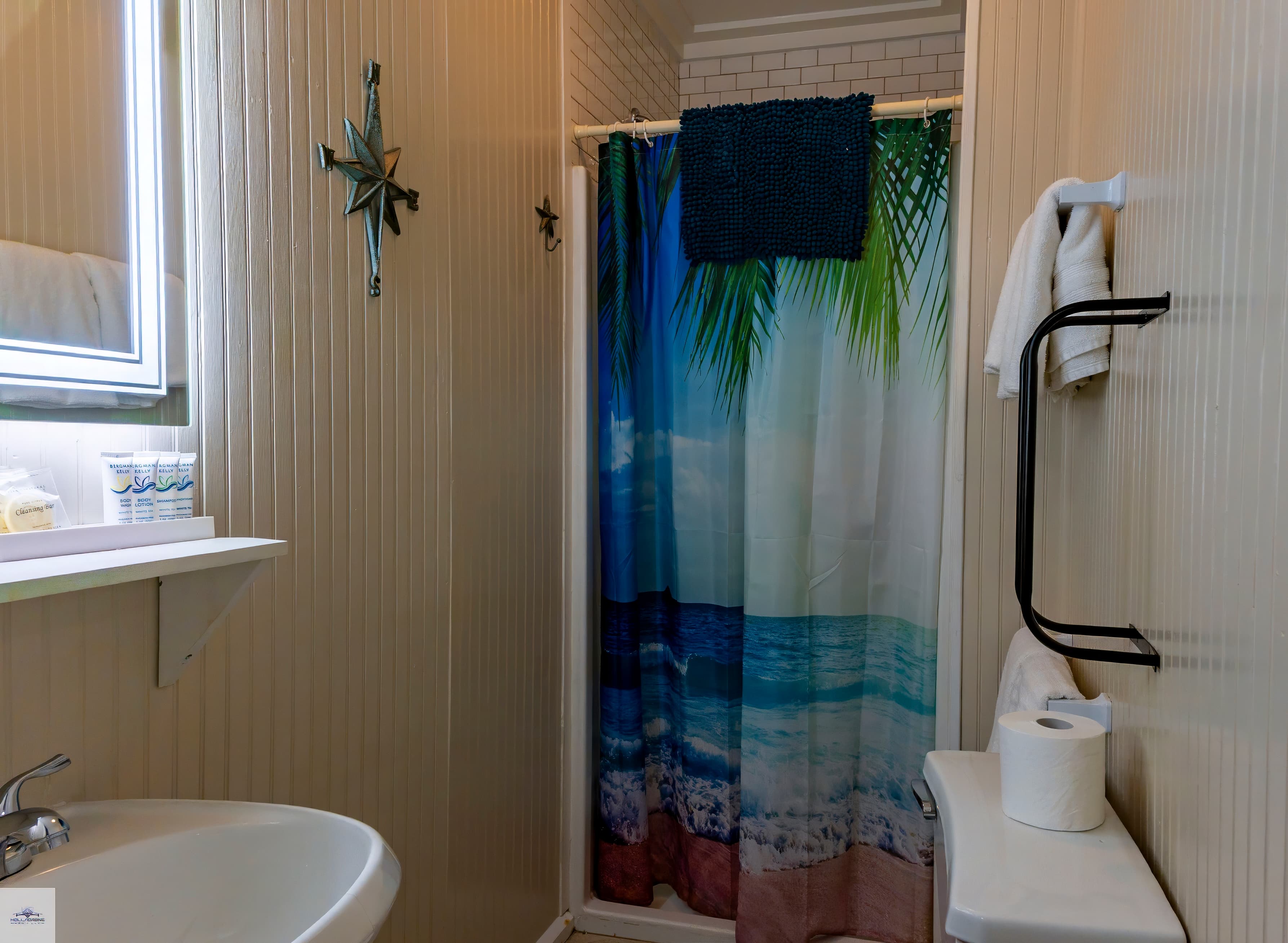 A compact bathroom featuring a sink, toilet, and a shower stall with a blue and tropical-themed curtain and light beige walls.