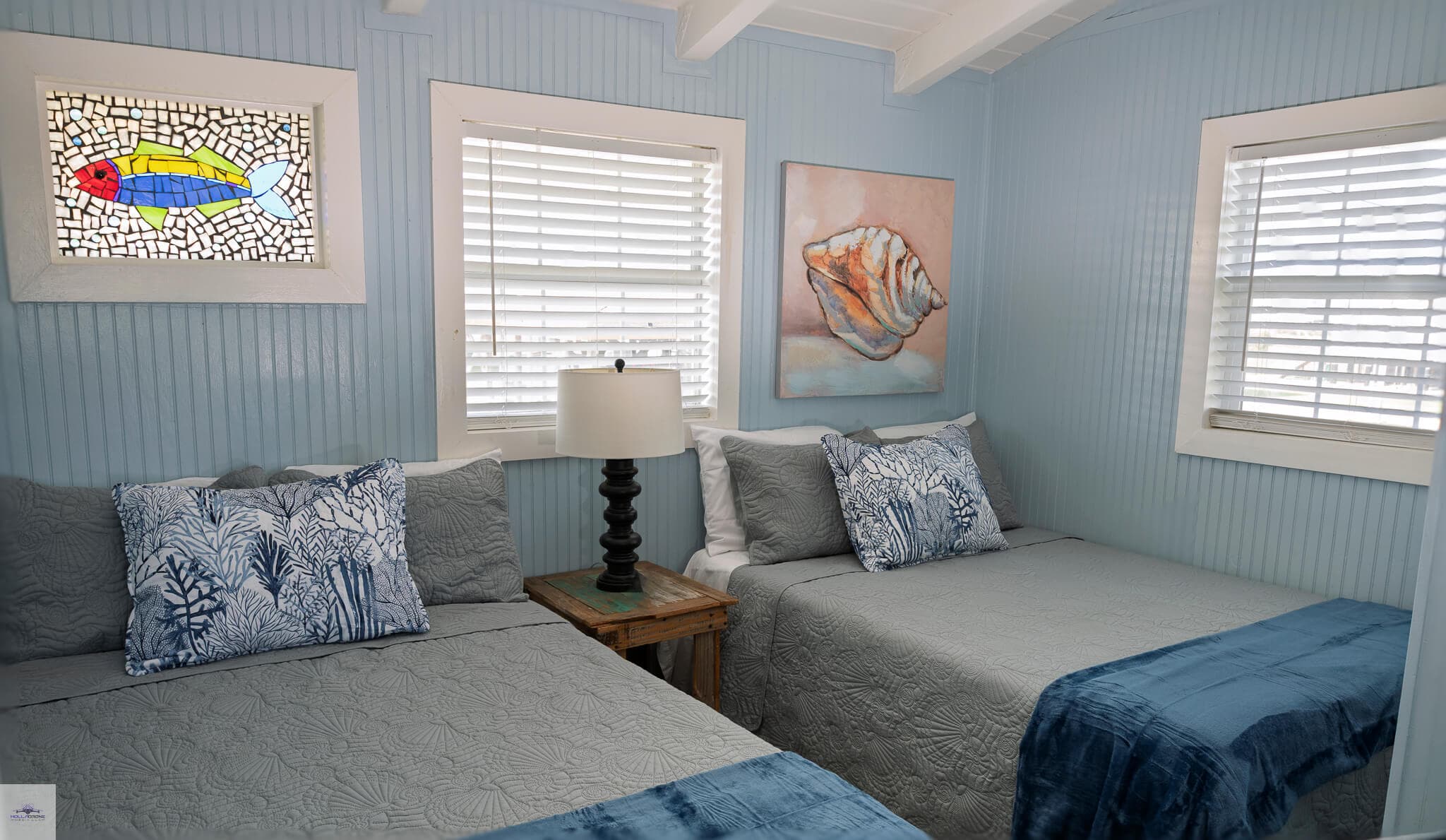 A cozy bedroom featuring two beds, a decorative lamp, and coastal-themed artwork on blue walls.