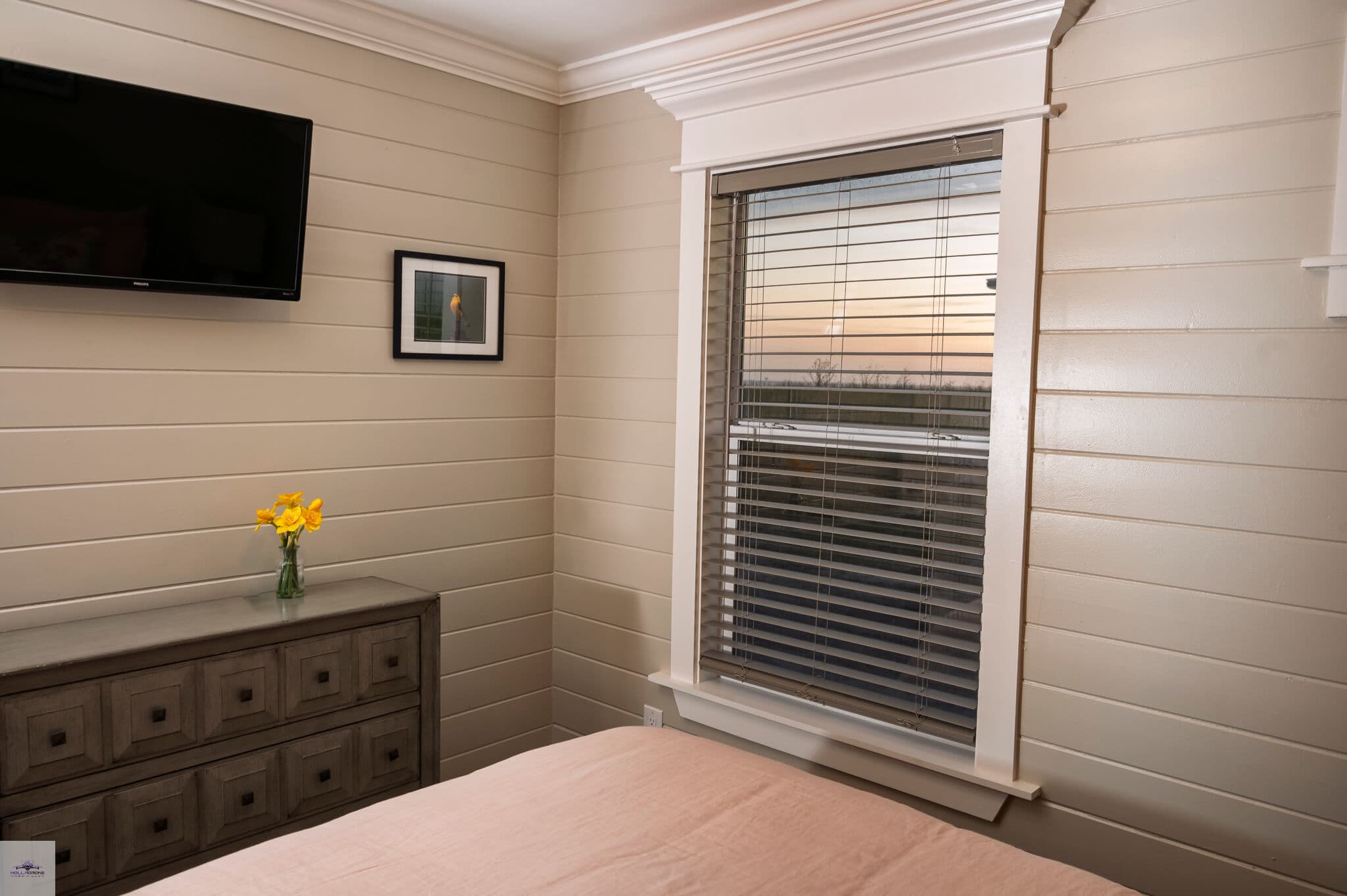 Cozy bedroom featuring a television, artwork, a dresser, and a window with a view of sunset.