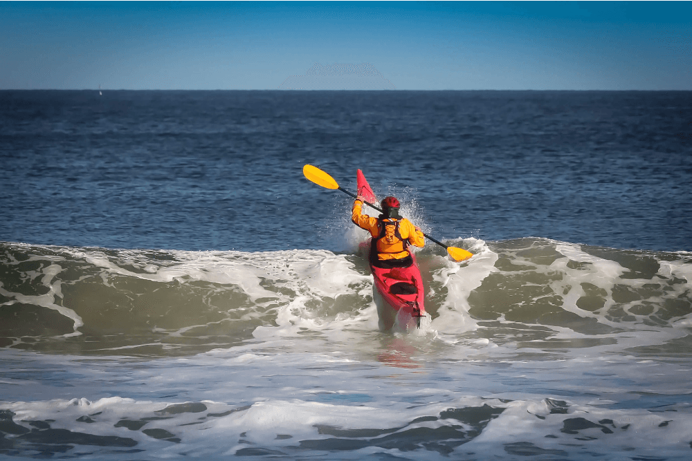 A kayaker paddles through ocean waves against a clear blue sky.