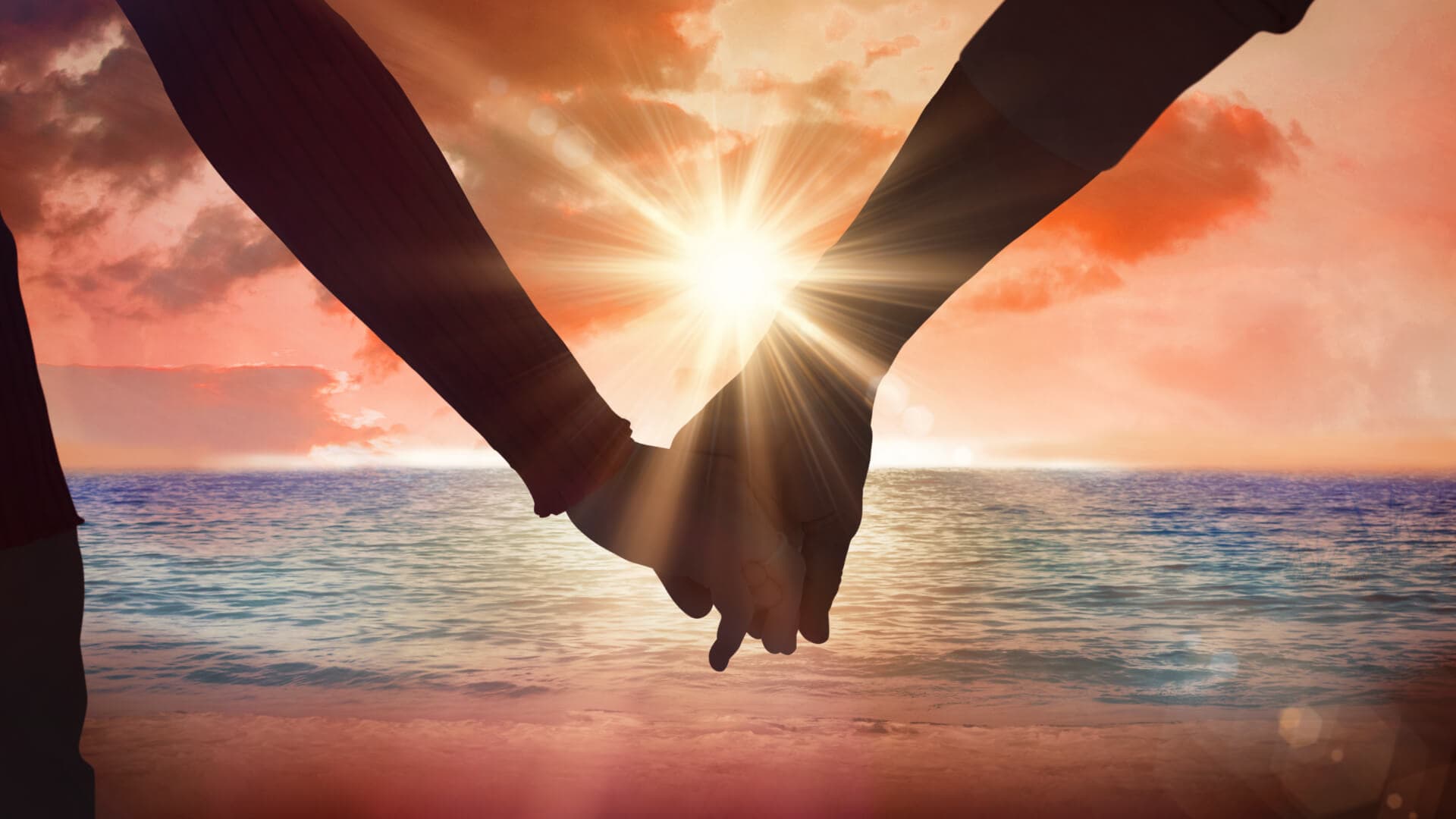 Two hands are intertwined against a scenic sunset over the ocean.