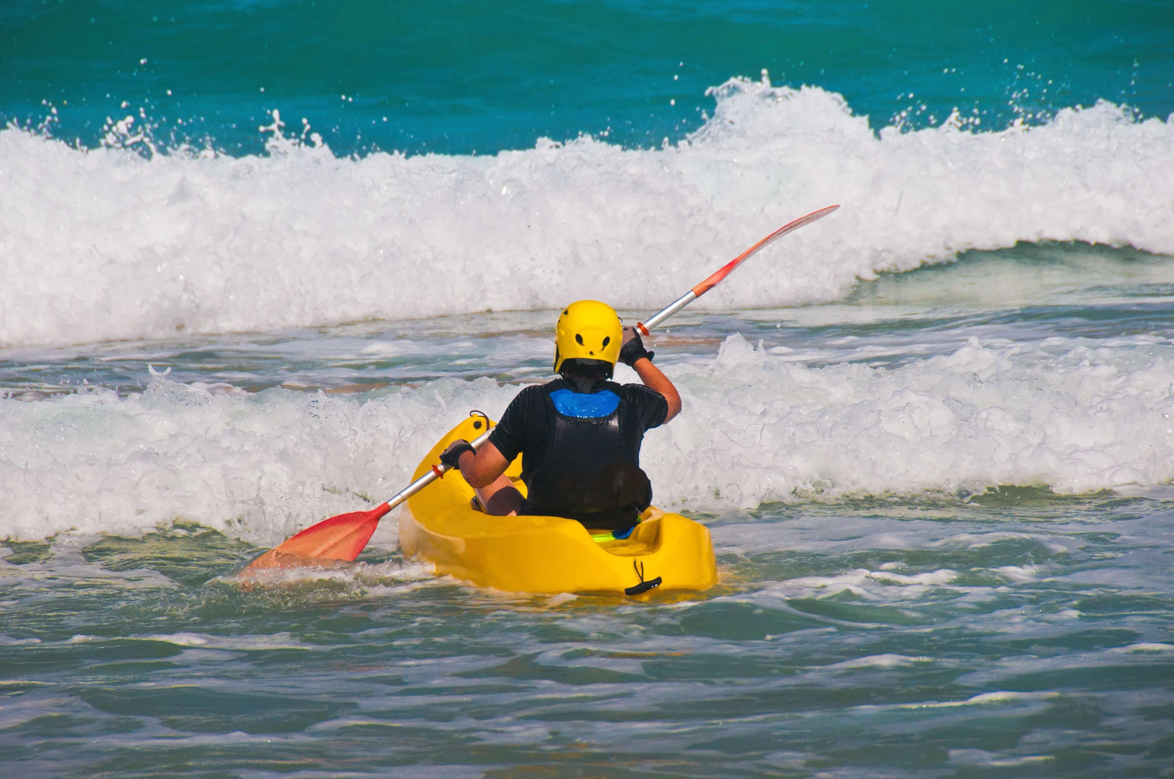 A person in a yellow kayak paddles through ocean waves wearing a helmet.