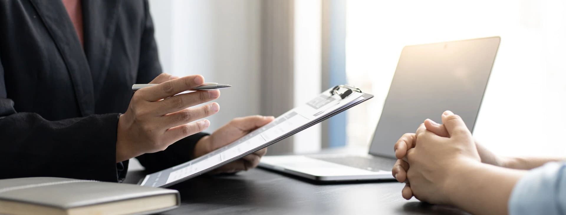 A person in a suit holds a clipboard while discussing with another individual at a desk with a laptop.